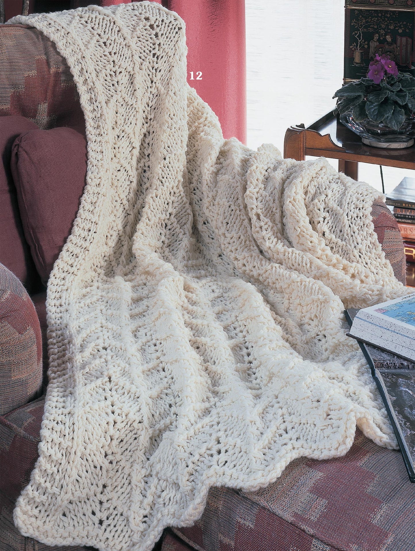 A cream-colored worsted weight knitted afghan from Leisure Arts' Big Book of Quick Knit Afghans, with a wavy textured pattern, is draped over an armchair by the window near pink curtains, a wooden table, plant, and books.