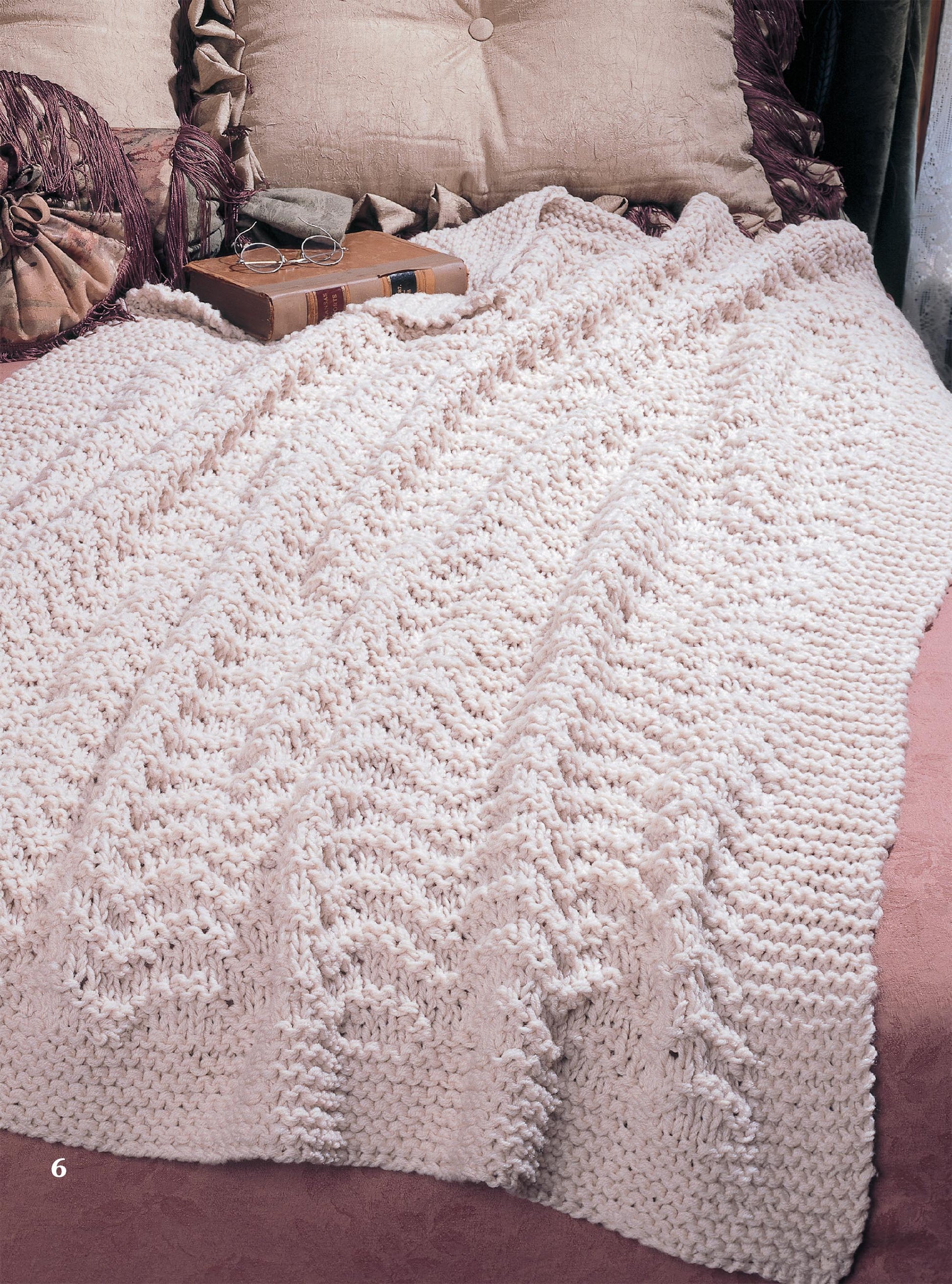 A knitted blanket resembling patterns from Leisure Arts’ Big Book of Quick Knit Afghans in light beige covers a bed with pink bedding and decorative pillows, one fringed and another with a bow. A book and glasses rest on the pillows.