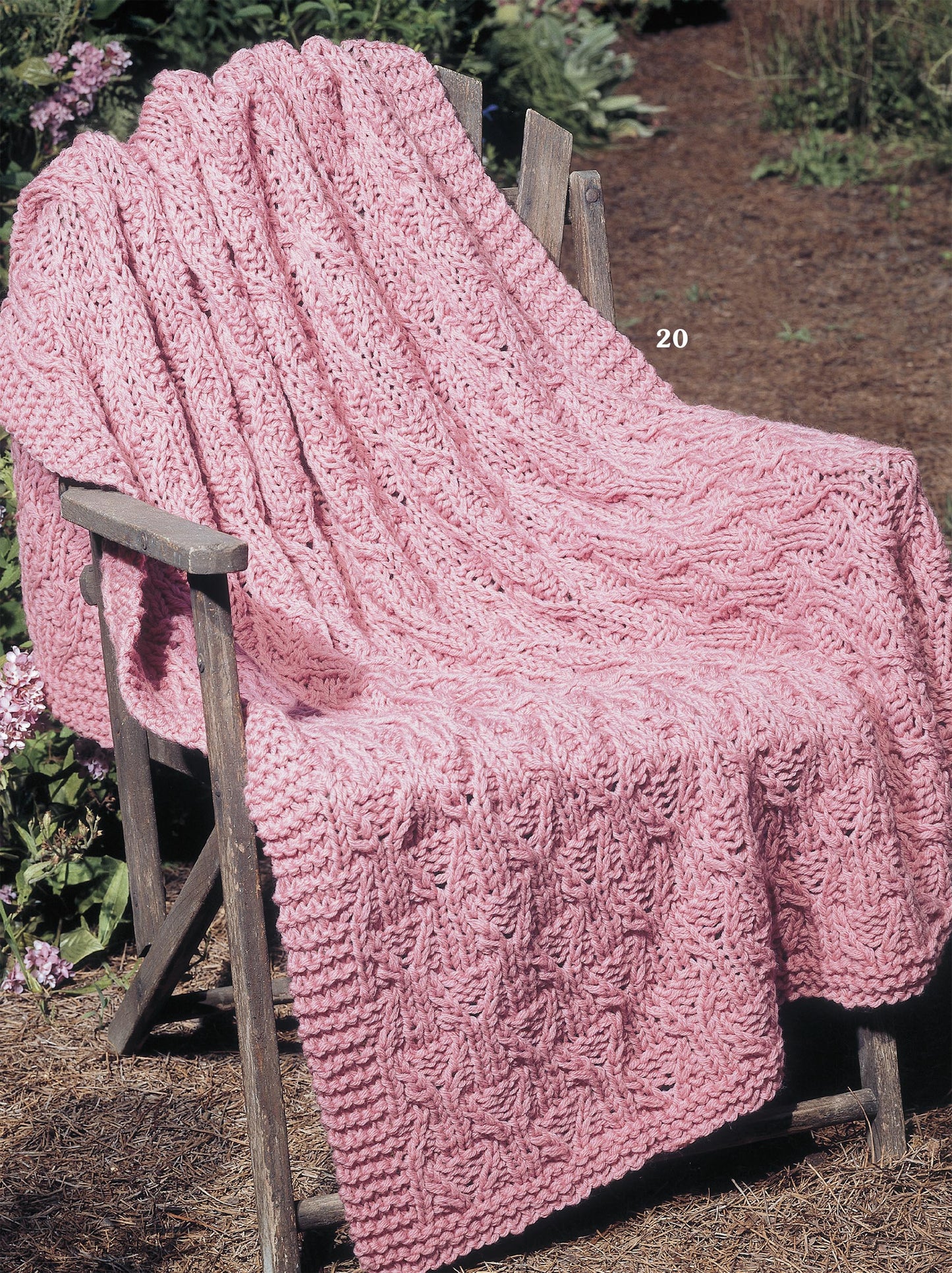 The pink, cable-patterned blanket, inspired by classic afghan styles from the Big Book of Quick Knit Afghans by Leisure Arts, is draped over a rustic wooden chair in a garden filled with greenery and soil.