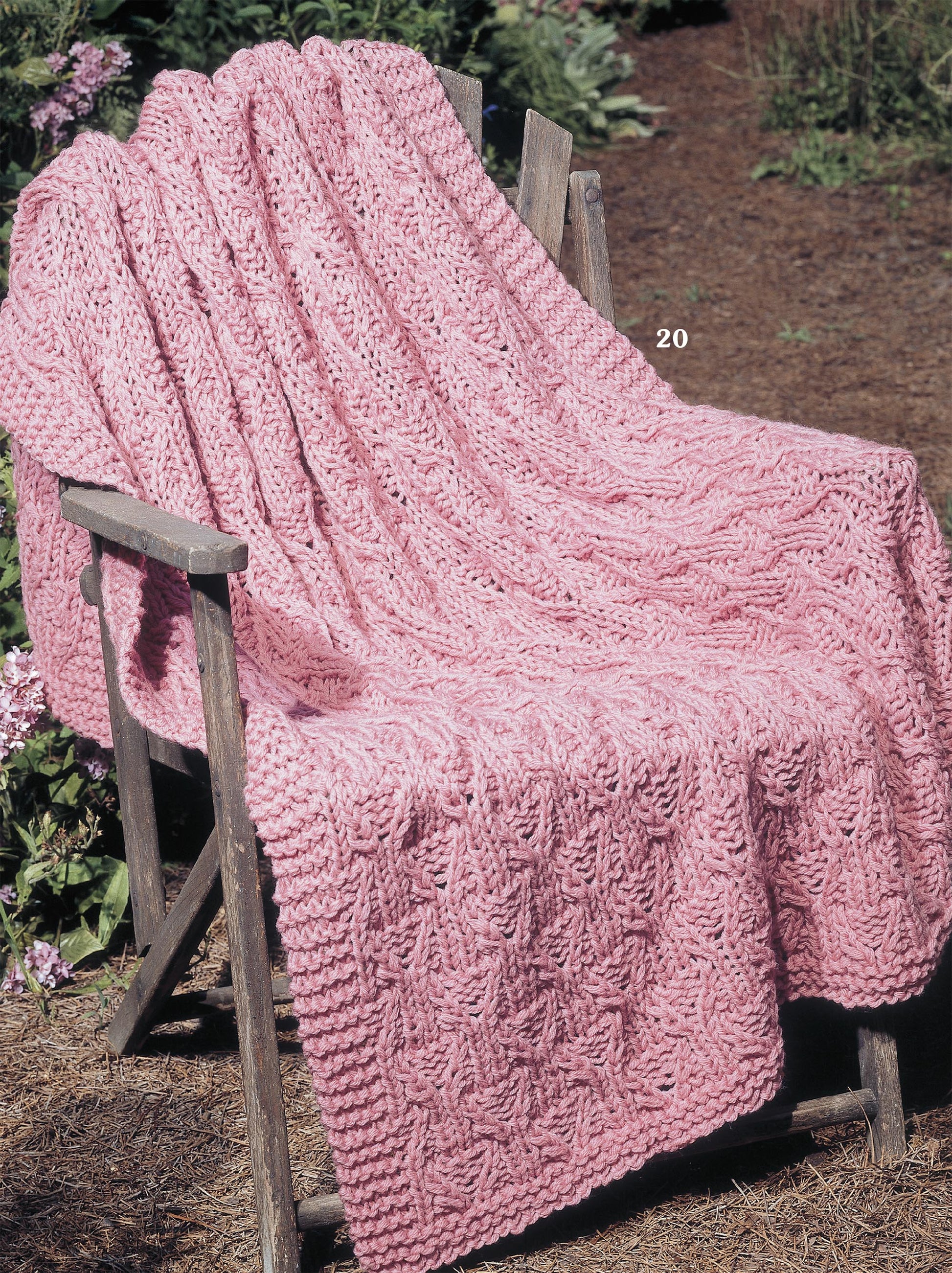 The pink, cable-patterned blanket, inspired by classic afghan styles from the Big Book of Quick Knit Afghans by Leisure Arts, is draped over a rustic wooden chair in a garden filled with greenery and soil.