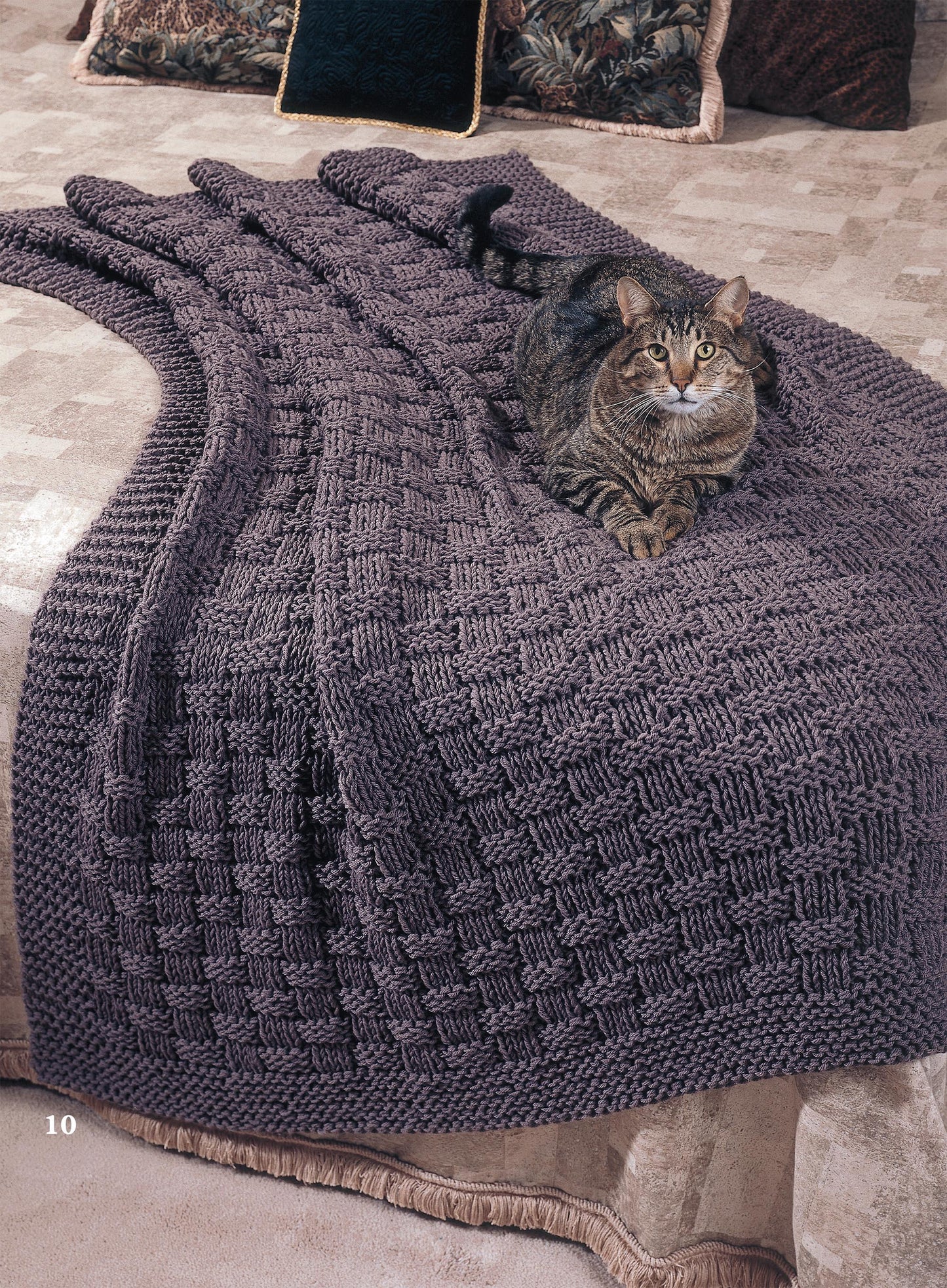 A tabby cat lounges on a dark purple afghan made with worsted weight yarn from Leisure Arts’ Big Book of Quick Knit Afghans, draped over a beige bed with patterned pillows arranged at the head.