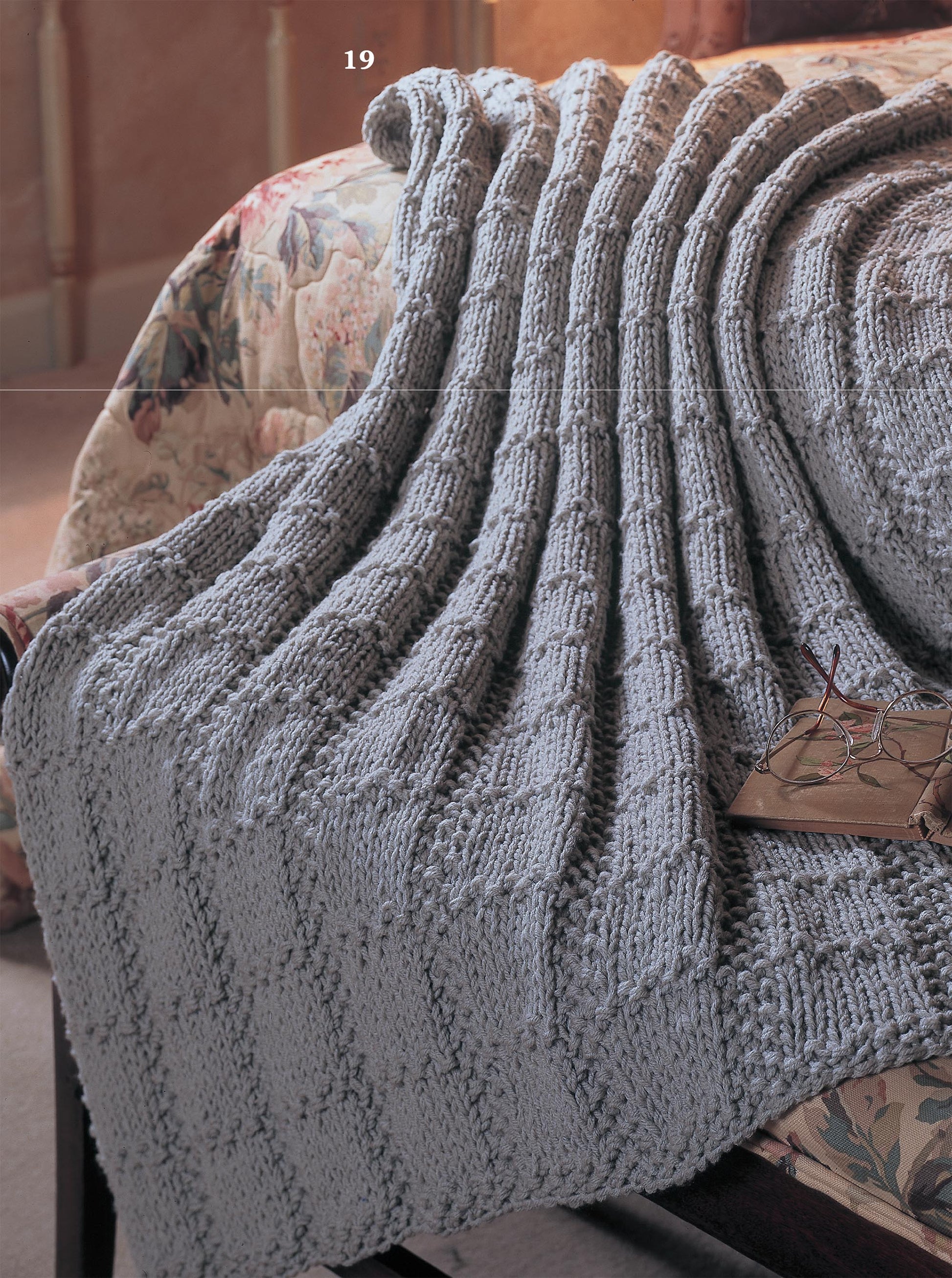 A cozy, light gray afghan from the "Big Book of Quick Knit Afghans" by Leisure Arts, featuring a checkered knit pattern, is draped over a floral bedspread with eyeglasses resting nearby.