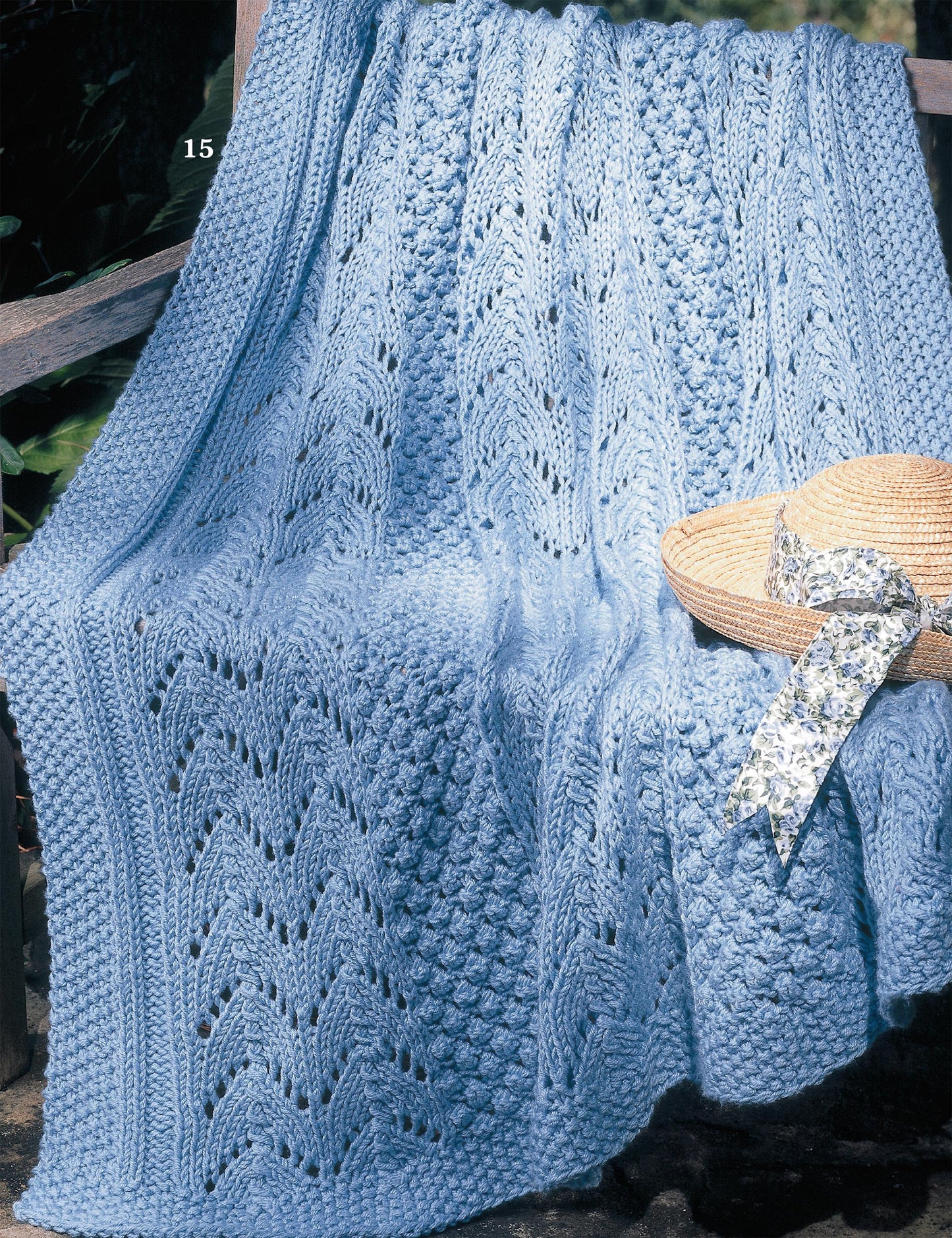 The "Big Book of Quick Knit Afghans" by Leisure Arts rests on a wooden bench, topped with a straw hat featuring a floral ribbon as sunlight streams over the cozy scene.