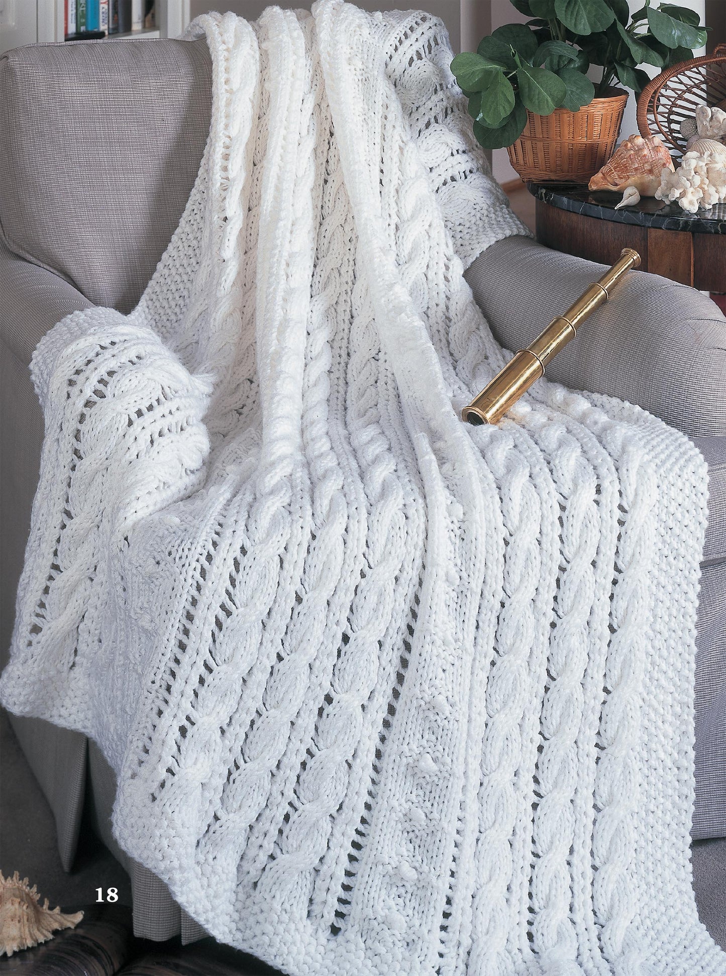 A cozy afghan from the Leisure Arts Big Book of Quick Knit Afghans drapes over a gray sofa arm, topped with a brass spyglass. Behind, leafy green plants and seashells decorate a wooden table.