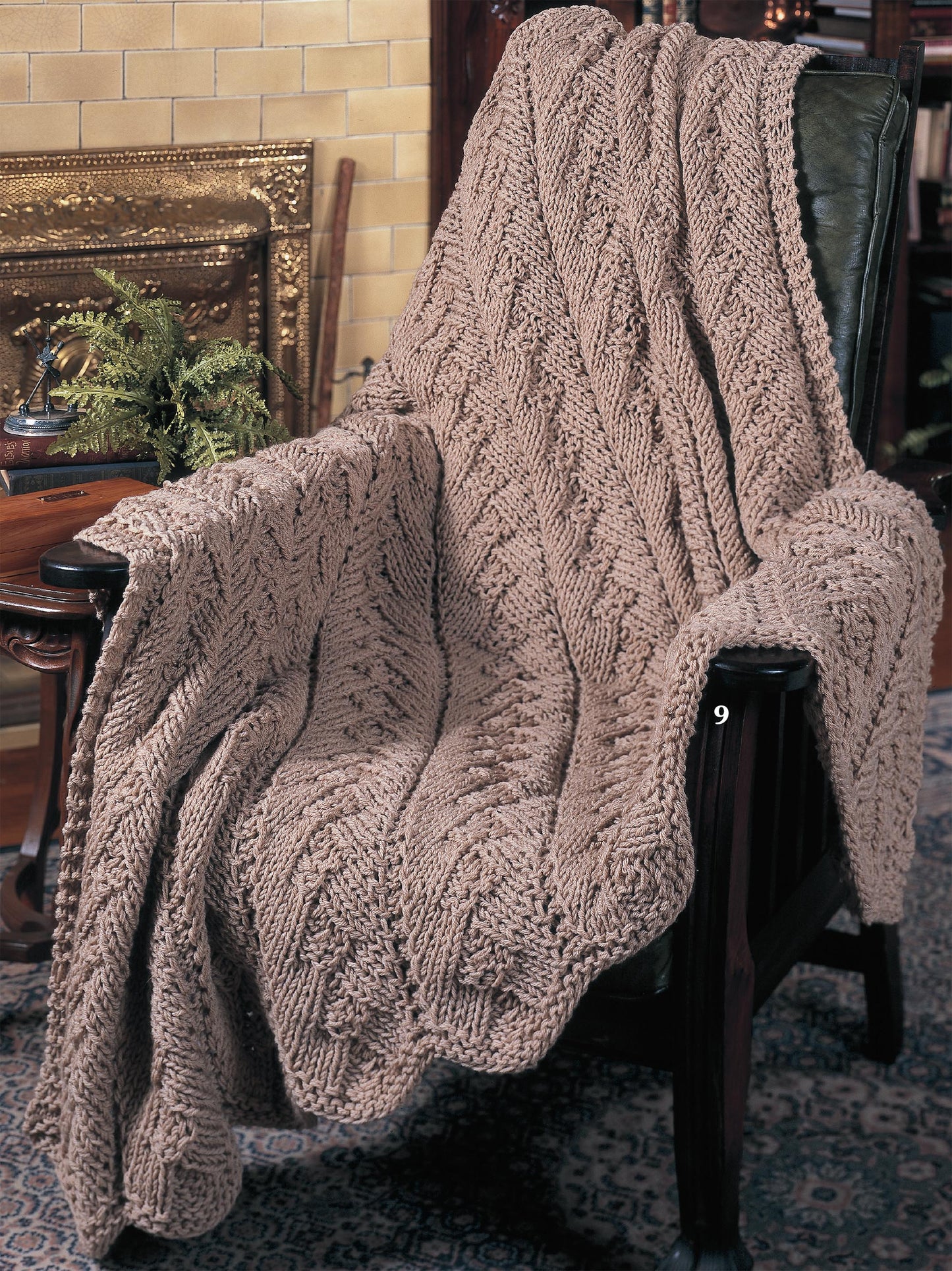 The Big Book of Quick Knit Afghans by Leisure Arts features cozy, worsted-weight, textured zigzag blankets in warm brown tones—perfect for draping over a chair in a fireside room with a patterned rug.