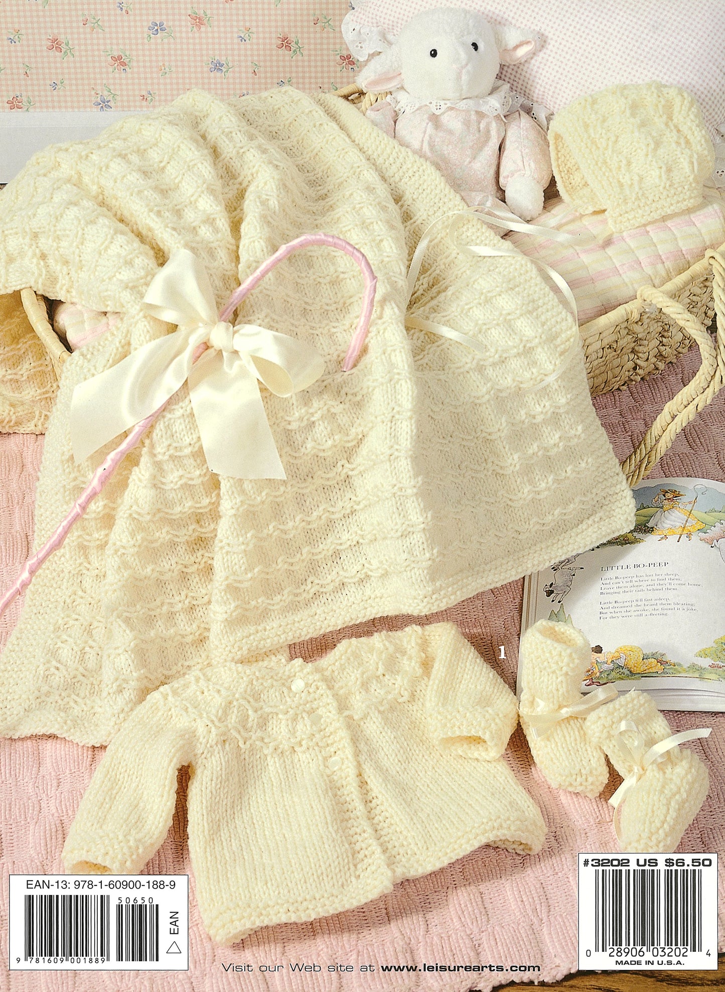 The Precious Layettes to Knit Digital Download by Leisure Arts features a cream-colored baby set with a blanket (bow detail), jacket, knit booties, and hat, shown on a pink quilt beside a plush lamb toy and an open book.