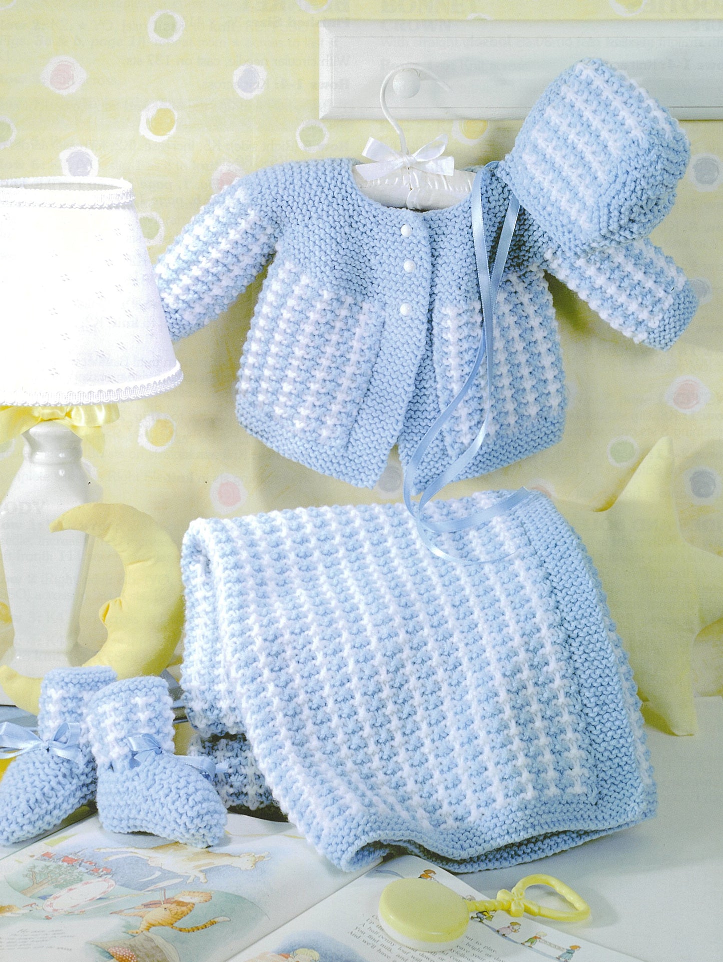 Precious Layettes to Knit" by Leisure Arts features a blue and white knit baby blanket with a matching layette—sweater (on hanger), hats, and booties—displayed near a lamp and baby decor against a soft yellow background.