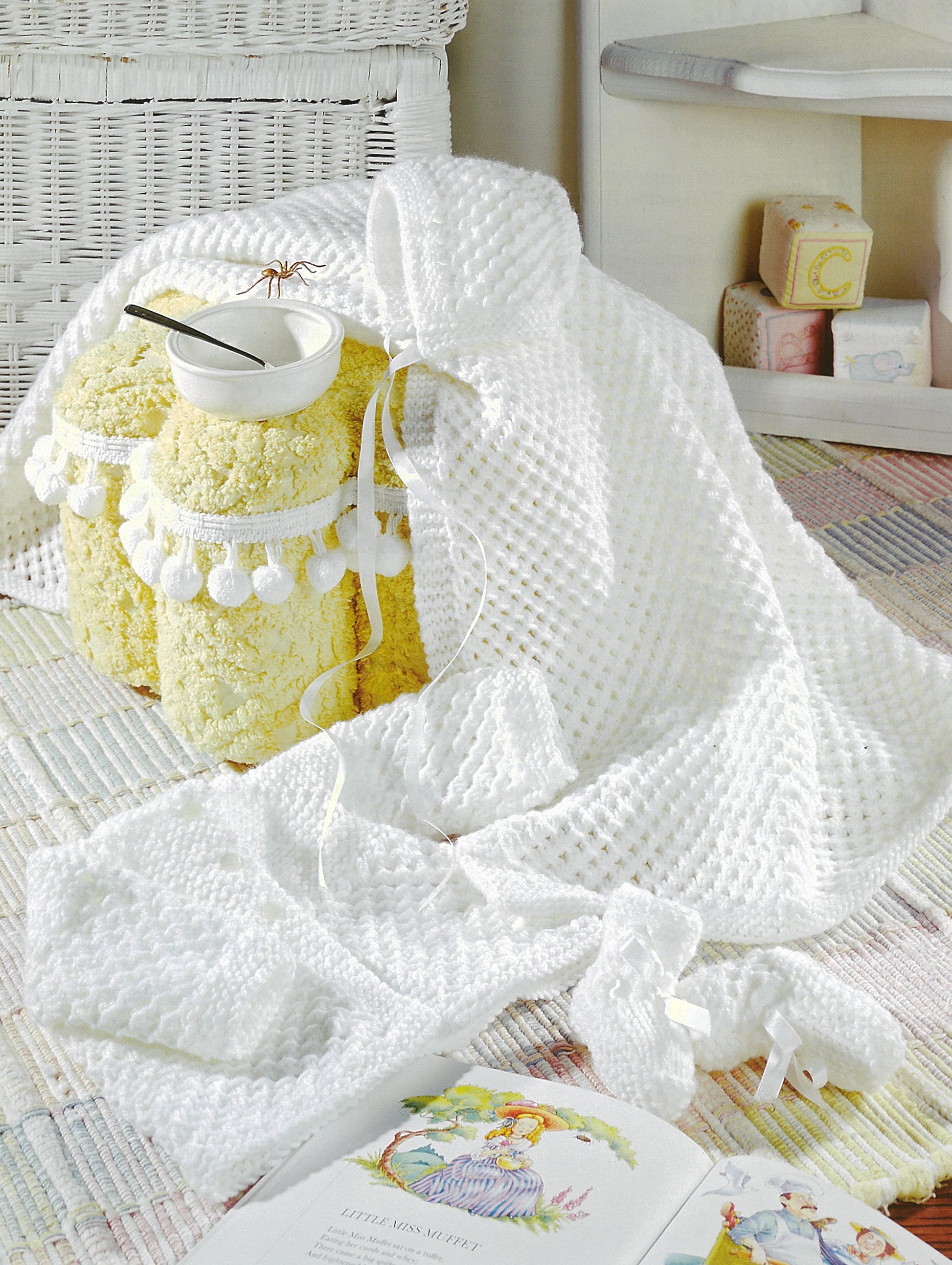 A cozy nursery features a soft baby blanket and matching hat from Leisure Arts’ “Precious Layettes to Knit,” draped over a yellow sheep-shaped stool with a bowl and spoon. Toy blocks and an open children’s book sit on a colorful rug nearby.