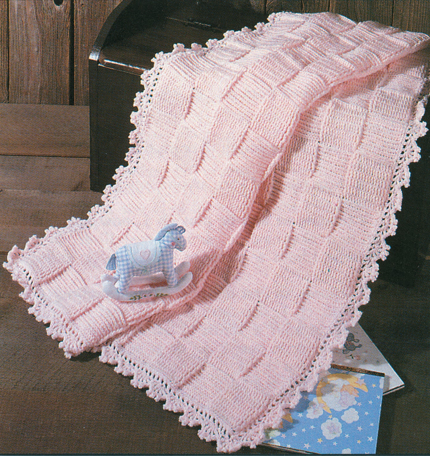 A soft pink knitted baby blanket, inspired by Snuggle-Up Baby Afghans—8 Projects from Leisure Arts, is draped over a wooden chest; a blue rocking horse toy and a children’s book rest nearby.