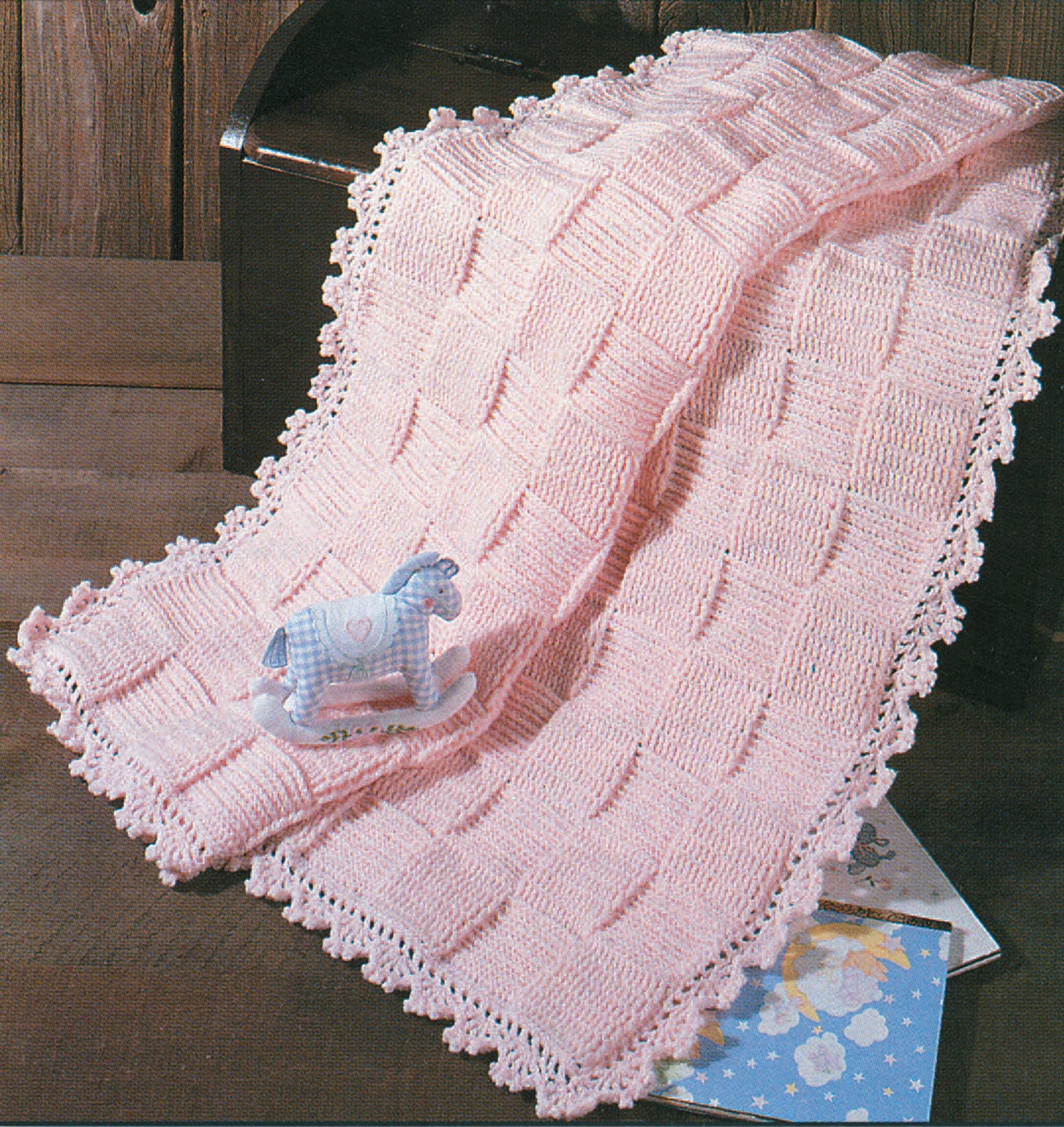 A soft pink knitted baby blanket, inspired by Snuggle-Up Baby Afghans—8 Projects from Leisure Arts, is draped over a wooden chest; a blue rocking horse toy and a children’s book rest nearby.