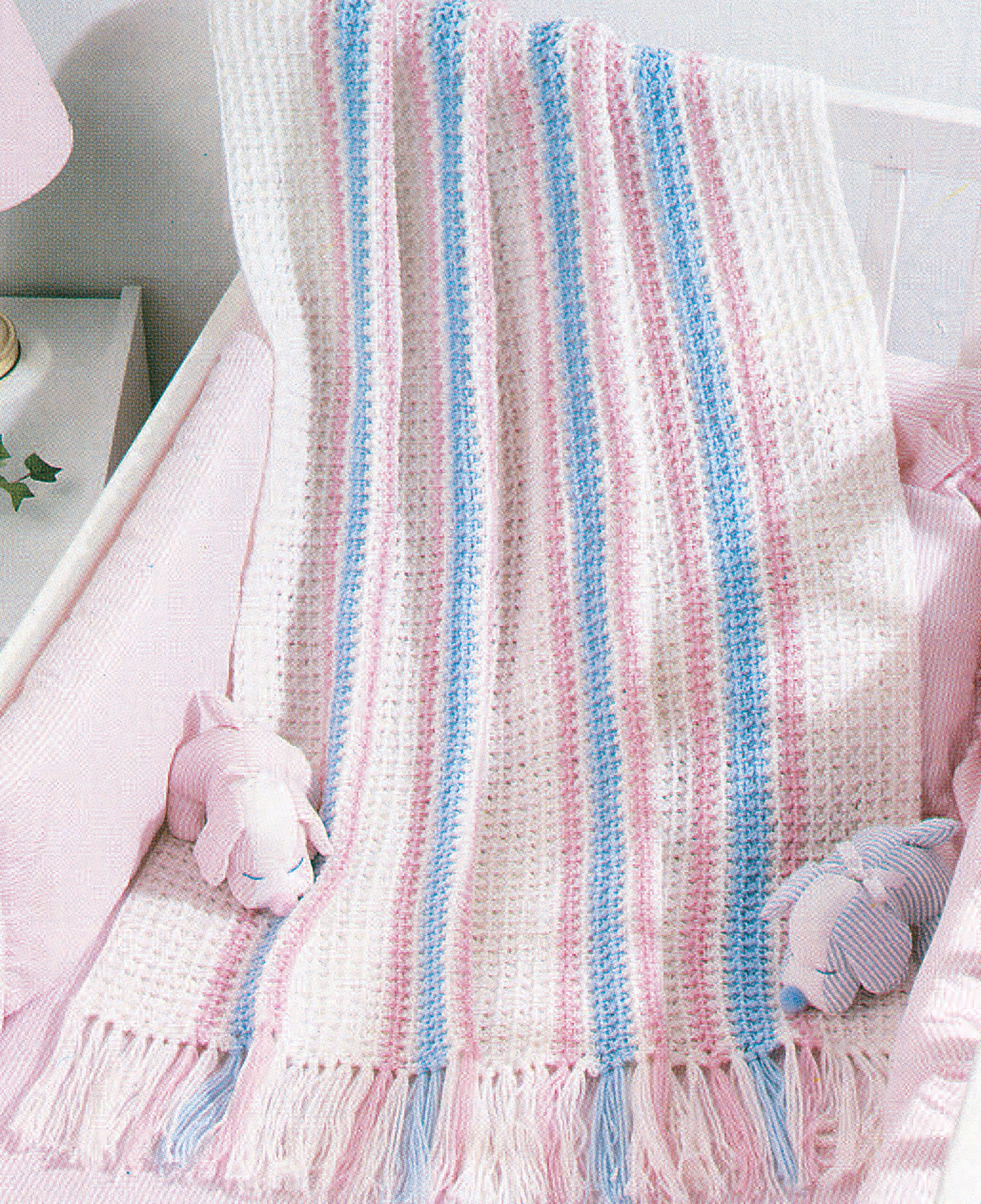 The Snuggle-Up Baby Afghans—8 Projects book by Leisure Arts features a beginner crochet baby blanket with pastel pink, blue, and white stripes plus fringe, shown draped over a crib beside plush toys—ideal for fans of baby afghans.
