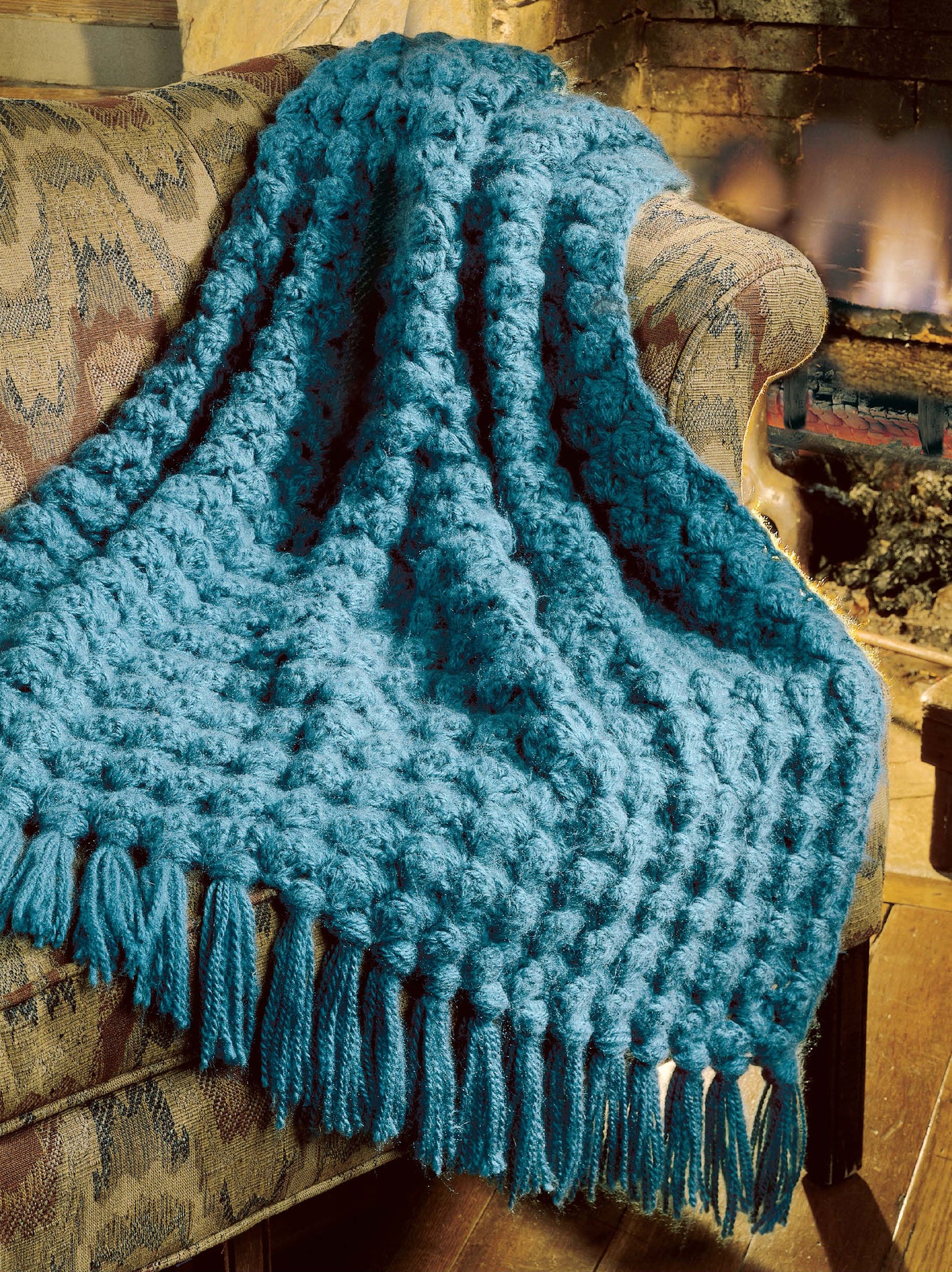 A blue, chunky crocheted afghan made using Leisure Arts' "Best of Terry Kimbrough Afghans Digital Download" is draped over a patterned couch by a fireplace, adding warmth and coziness to the room.