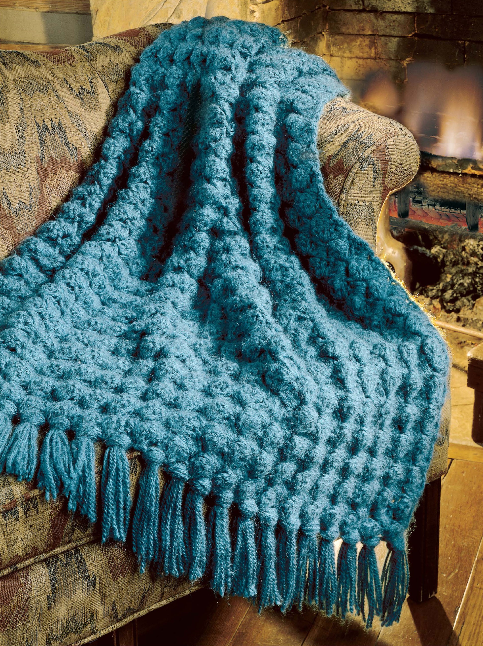 A blue, chunky crocheted afghan made using Leisure Arts' "Best of Terry Kimbrough Afghans Digital Download" is draped over a patterned couch by a fireplace, adding warmth and coziness to the room.