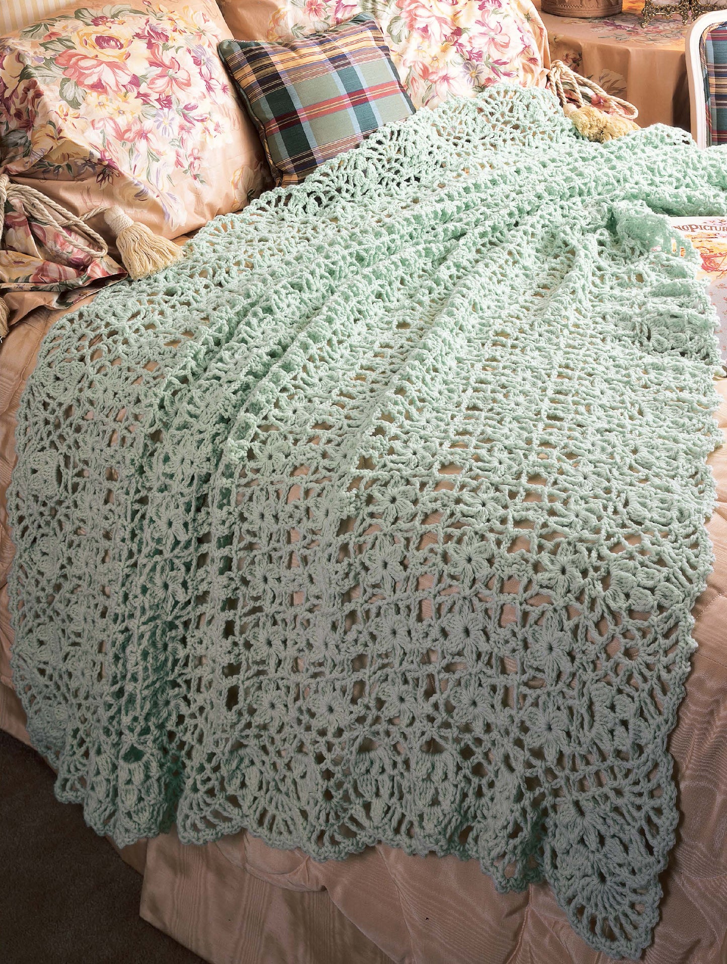 A light blue crocheted blanket from "Best of Terry Kimbrough Afghans" by Leisure Arts, featuring a Victorian lace-inspired pattern, is draped over a bed with floral and plaid pillows and a cream quilted bedspread.