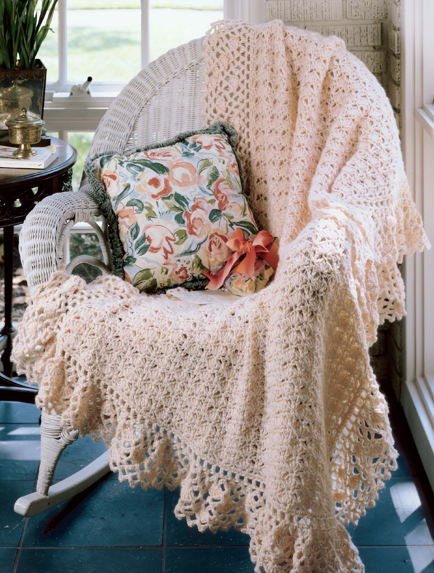 A white wicker chair is adorned with the "Best of Terry Kimbrough Afghans" by Leisure Arts and a floral pillow, placed by a sunlit window.