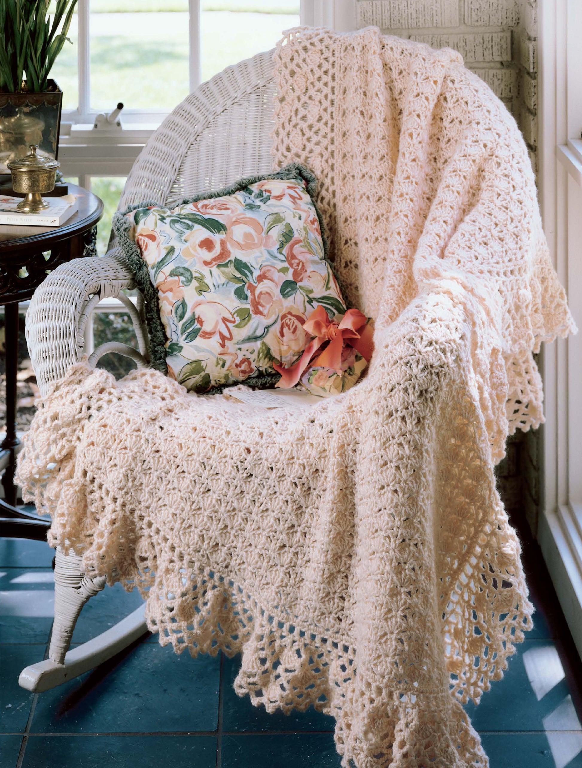 A white wicker chair is adorned with the "Best of Terry Kimbrough Afghans" by Leisure Arts and a floral pillow, placed by a sunlit window.