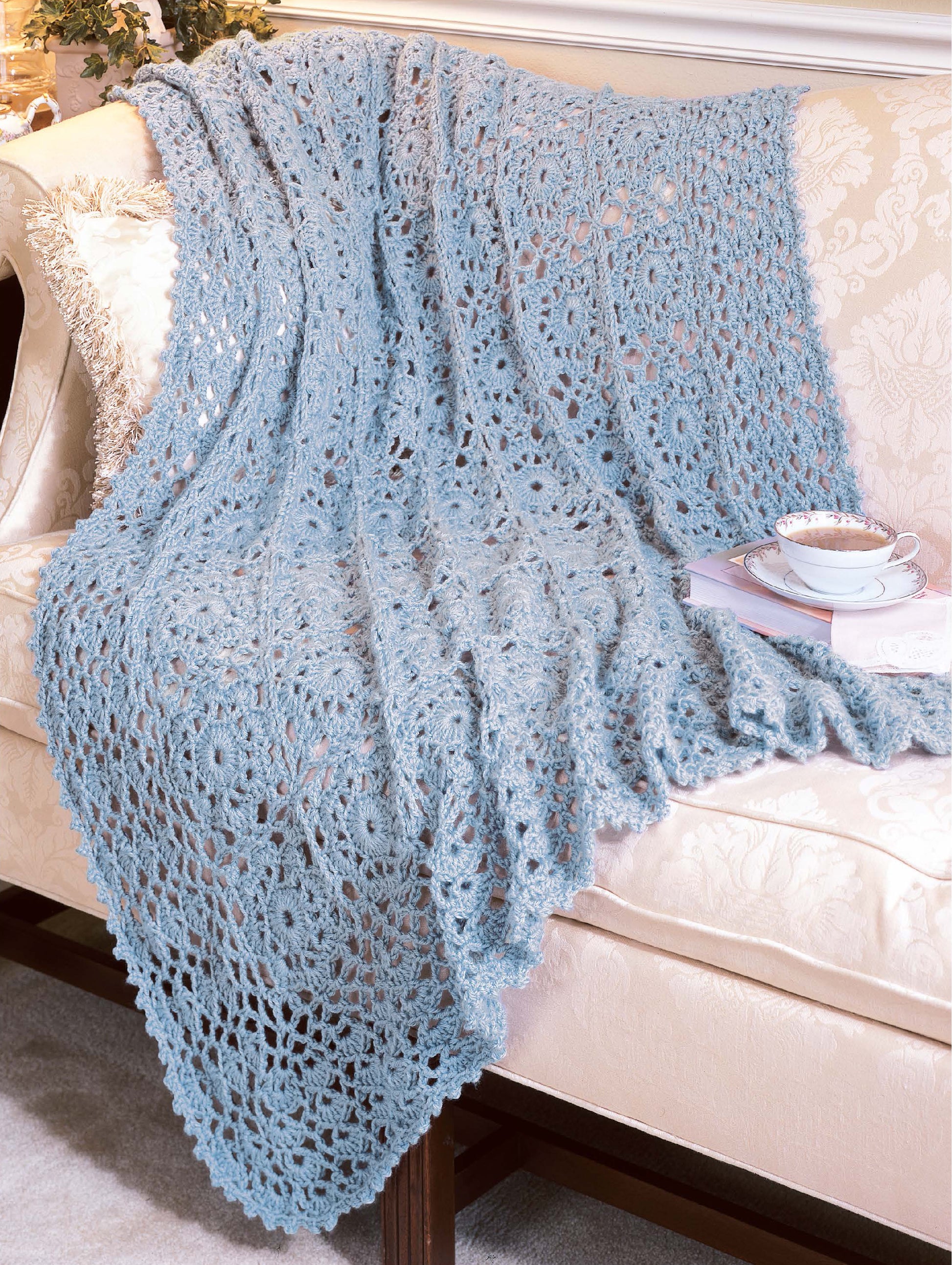 A soft blue crocheted blanket from the "Best of Terry Kimbrough Afghans" by Leisure Arts is draped over a cream floral sofa, with a cup and saucer on the arm—bringing cozy Victorian-inspired charm to your living room.