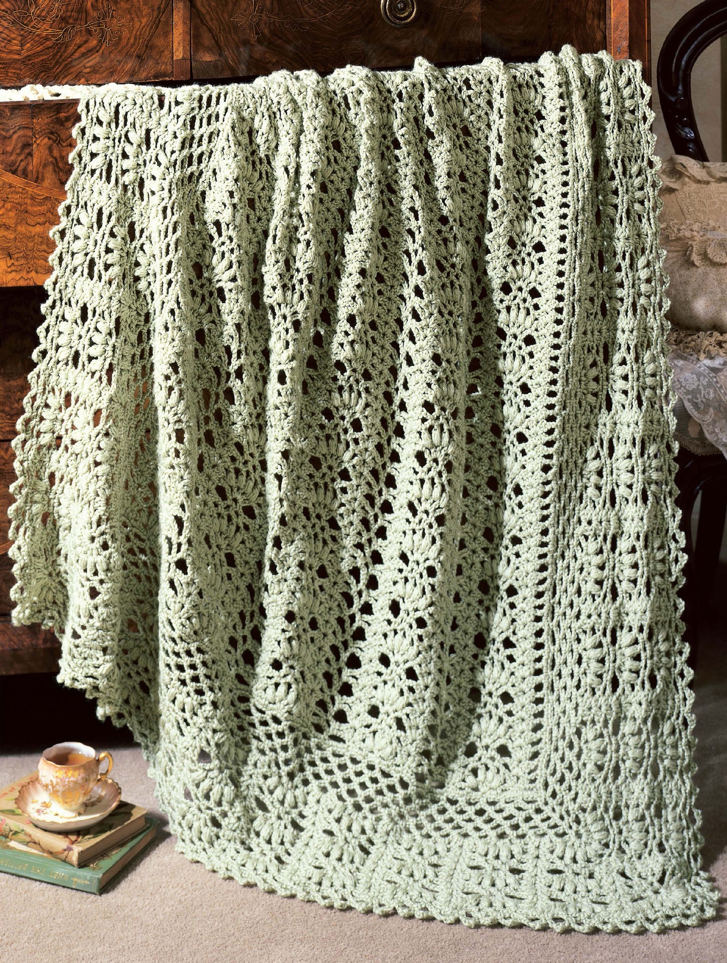 A light green, lacy crocheted afghan from "Best of Terry Kimbrough Afghans" by Leisure Arts drapes over a wooden table, with a teacup and book nearby, creating a cozy, vintage vibe.