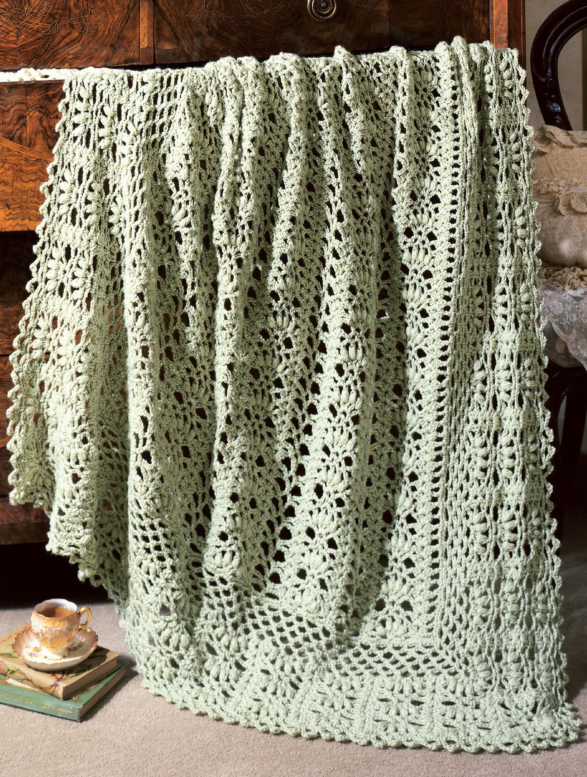 A light green, lacy crocheted afghan from "Best of Terry Kimbrough Afghans" by Leisure Arts drapes over a wooden table, with a teacup and book nearby, creating a cozy, vintage vibe.