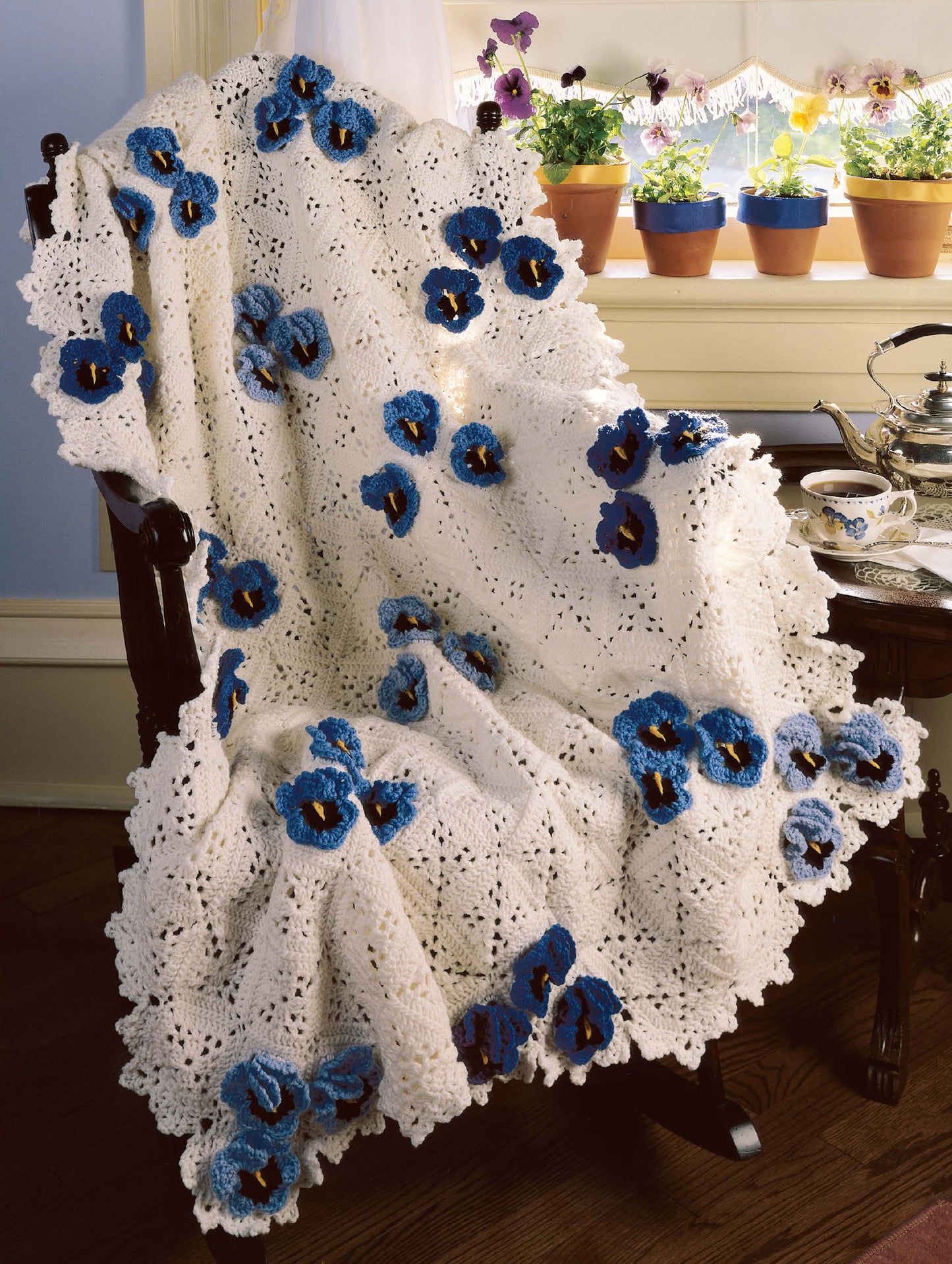 The Best of Terry Kimbrough Afghans by Leisure Arts, a cream-colored crocheted blanket with blue and purple flower accents and Victorian lace-inspired finishing, is draped over a dark wooden chair; potted flowers and a teapot are on the windowsill.