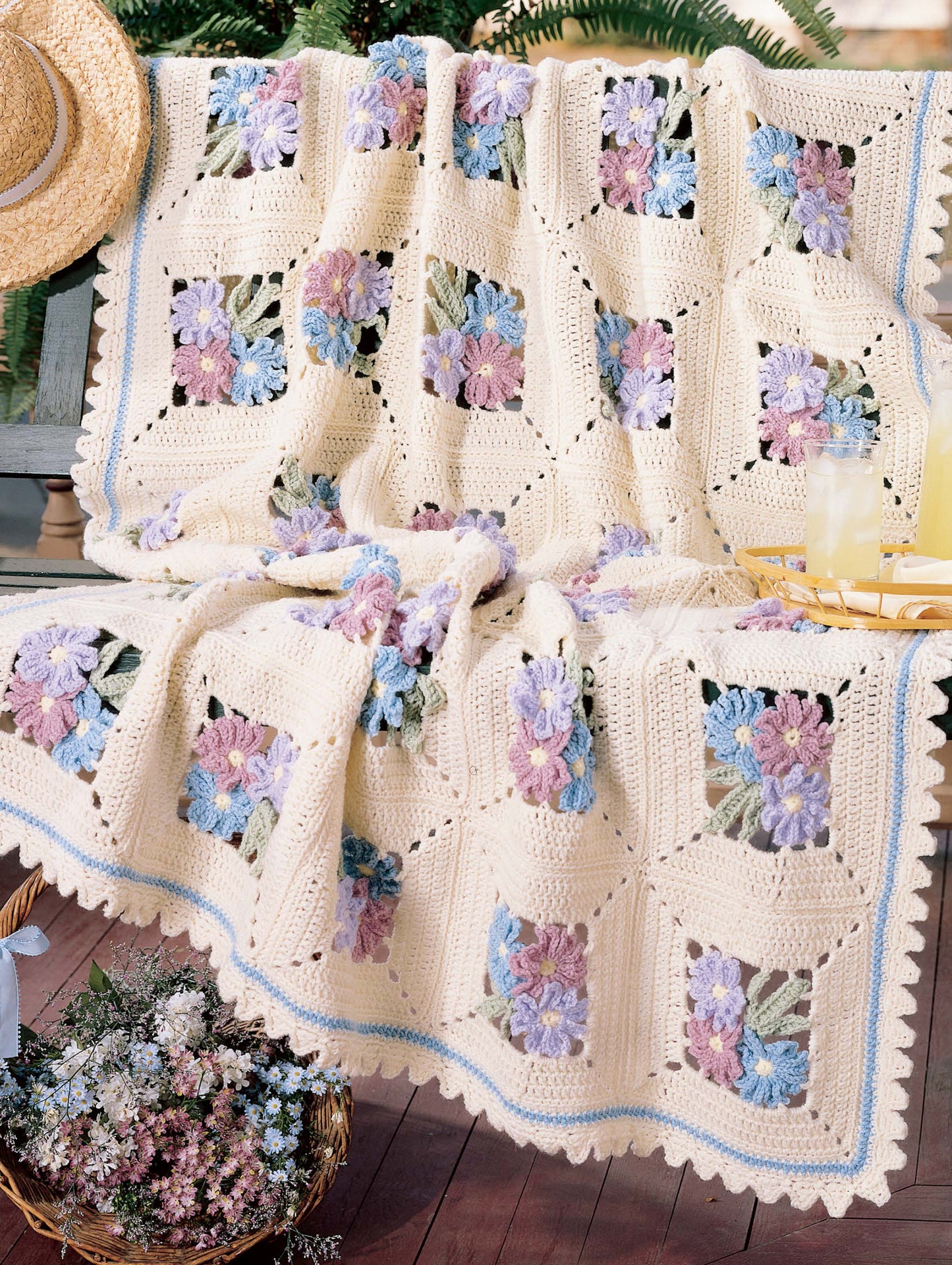 The "Best of Terry Kimbrough Afghans" by Leisure Arts, featuring a cream crocheted lace design with purple, blue, and pink flowers, is draped over a bench, paired with a straw hat, lemonade glasses, and flowers for a cozy summer look.