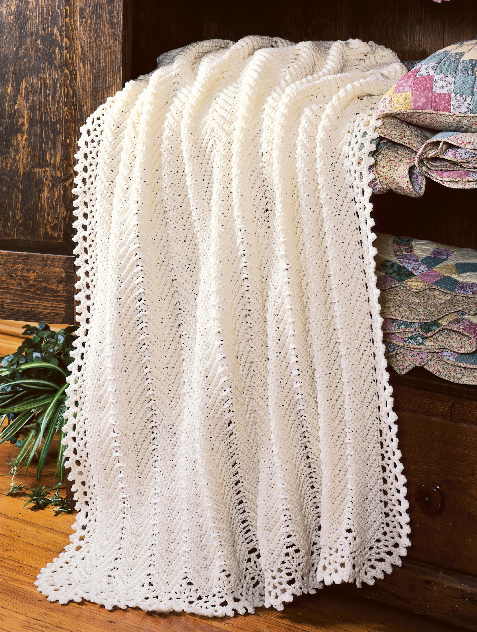 A cream crocheted blanket from "Best of Terry Kimbrough Afghans" by Leisure Arts drapes over a wooden shelf beside a colorful patchwork quilt, while a green potted plant sits on the floor nearby.