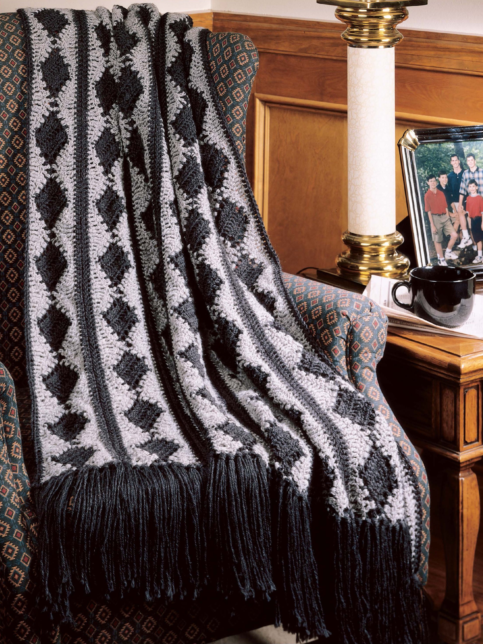 The Best of Terry Kimbrough Afghans by Leisure Arts, featuring a black and gray diamond pattern with fringe and elegant finishing, is draped over an upholstered armchair beside a wooden table holding a lamp, photo frame, and mug.