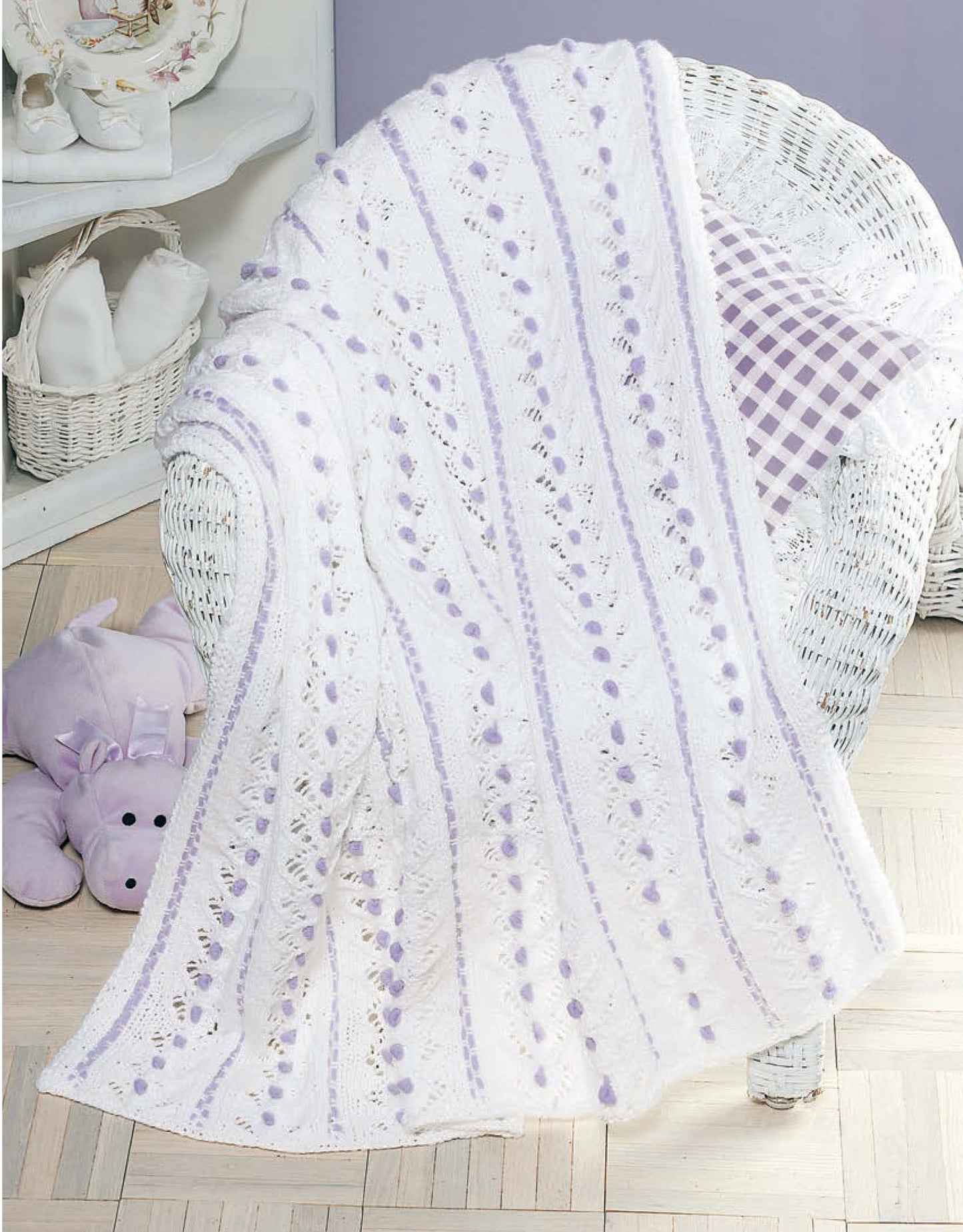 A white wicker chair displays a white and lavender afghan from Leisure Arts' Our Best Knit Baby Afghans, beside a purple gingham pillow. A plush purple hippo toy is nearby; white shelves with folded towels are in the background.