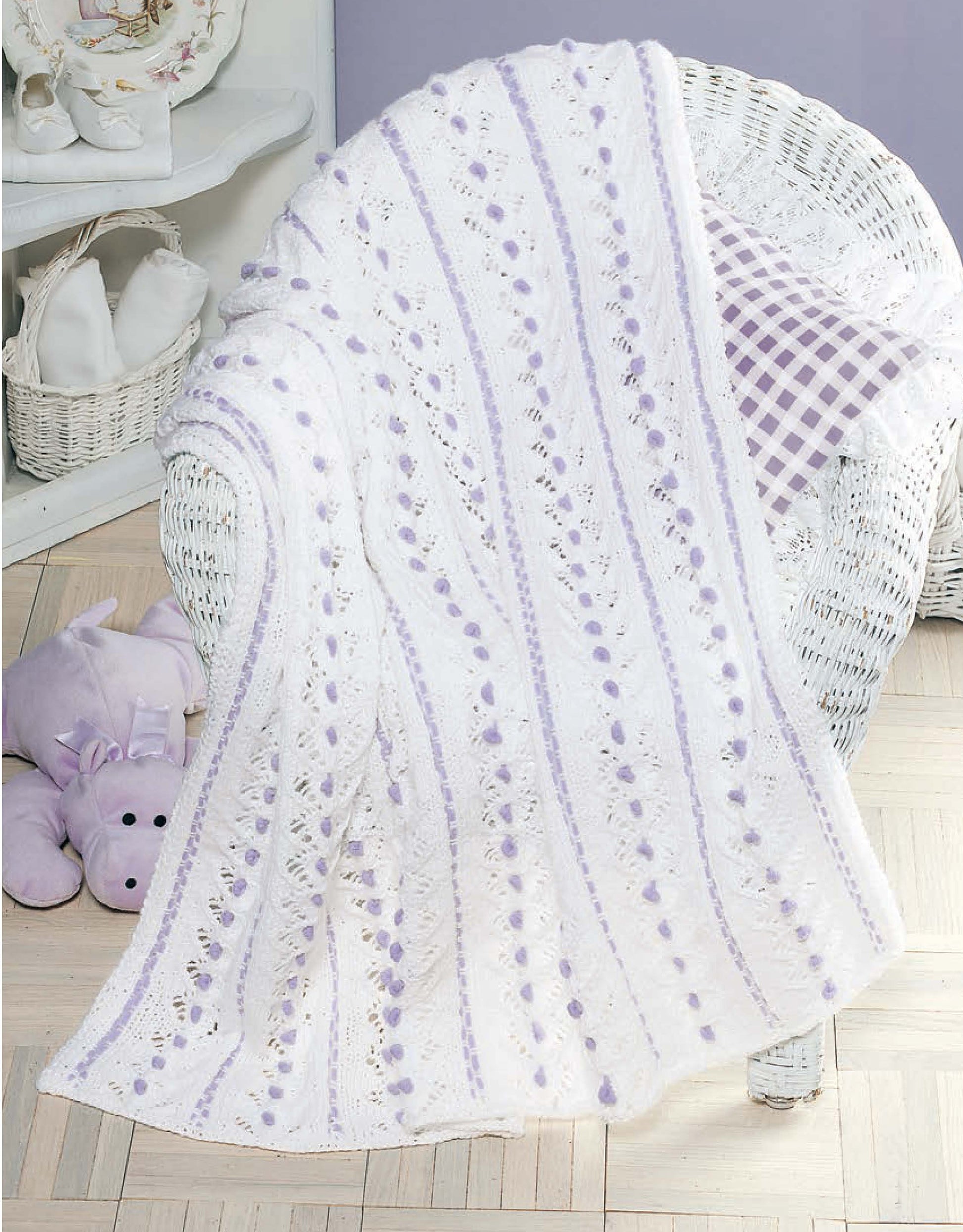 A white wicker chair displays a white and lavender afghan from Leisure Arts' Our Best Knit Baby Afghans, beside a purple gingham pillow. A plush purple hippo toy is nearby; white shelves with folded towels are in the background.