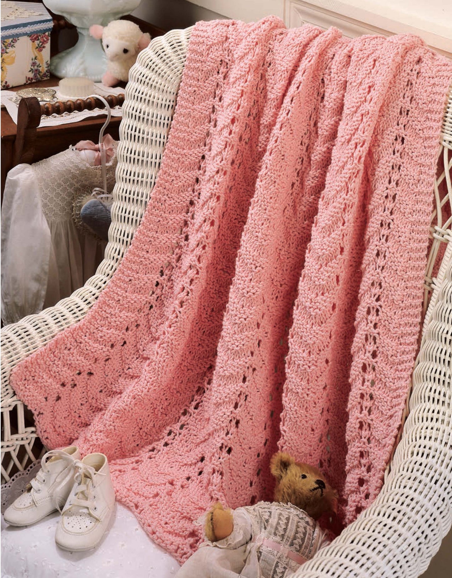 A pink blanket from Leisure Arts' "Our Best Knit Baby Afghans" is draped over a white wicker chair with a teddy bear, baby shoes, and soft toys nearby in a cozy vintage-style nursery.