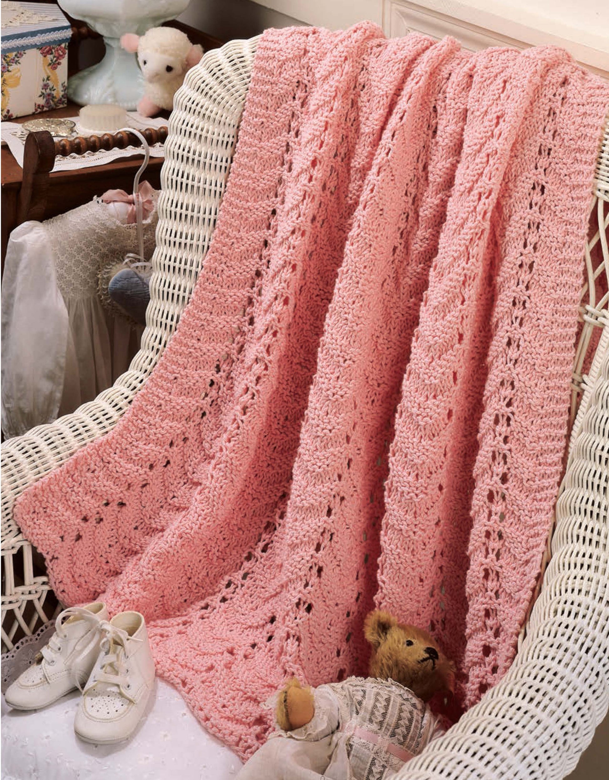A pink blanket from Leisure Arts' "Our Best Knit Baby Afghans" is draped over a white wicker chair with a teddy bear, baby shoes, and soft toys nearby in a cozy vintage-style nursery.