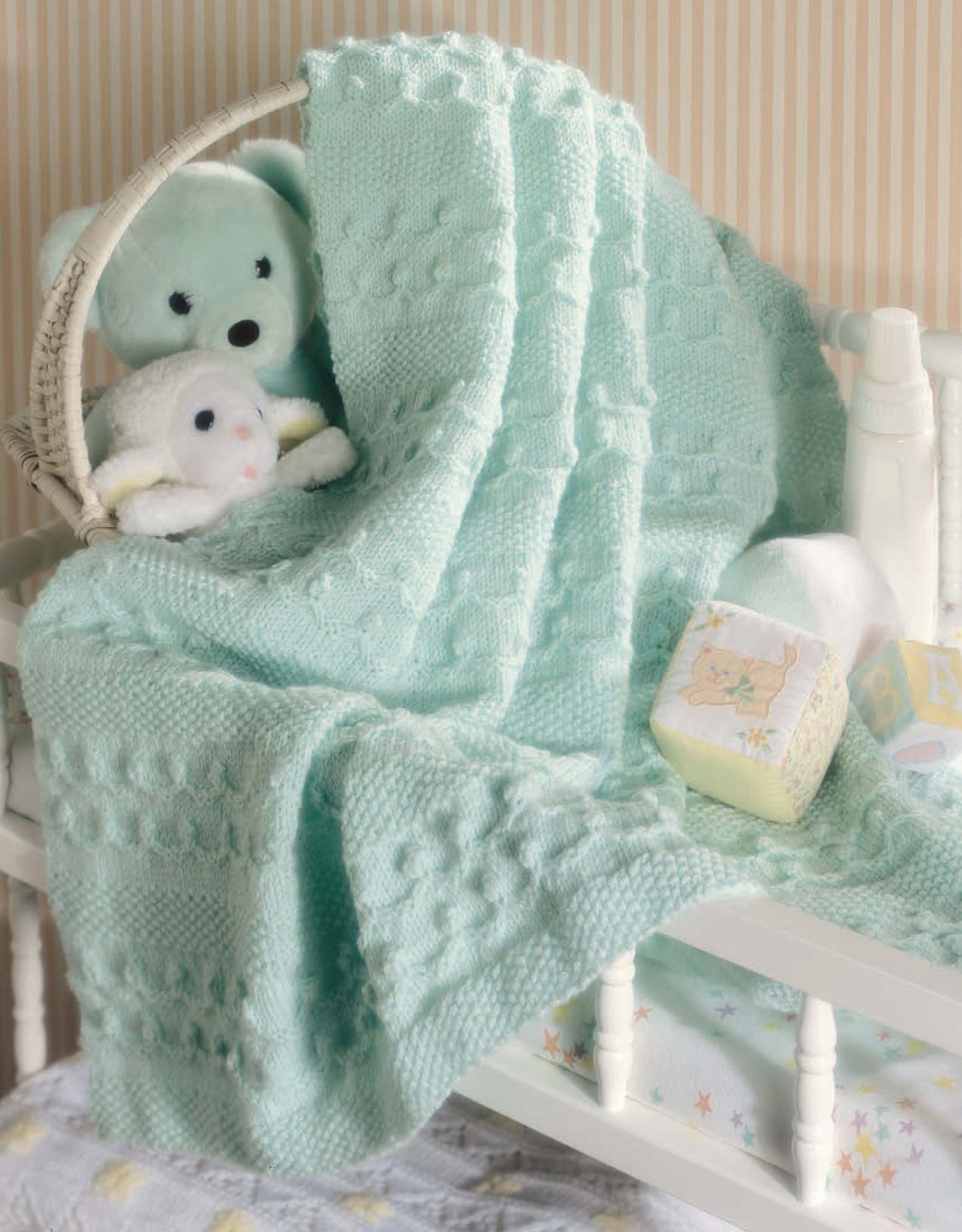 A soft mint green baby blanket from "Our Best Knit Baby Afghans" by Leisure Arts is draped over a white crib with a plush bear, lamb, wicker basket, pastel alphabet blocks, baby bottle, and striped wallpaper in the background.