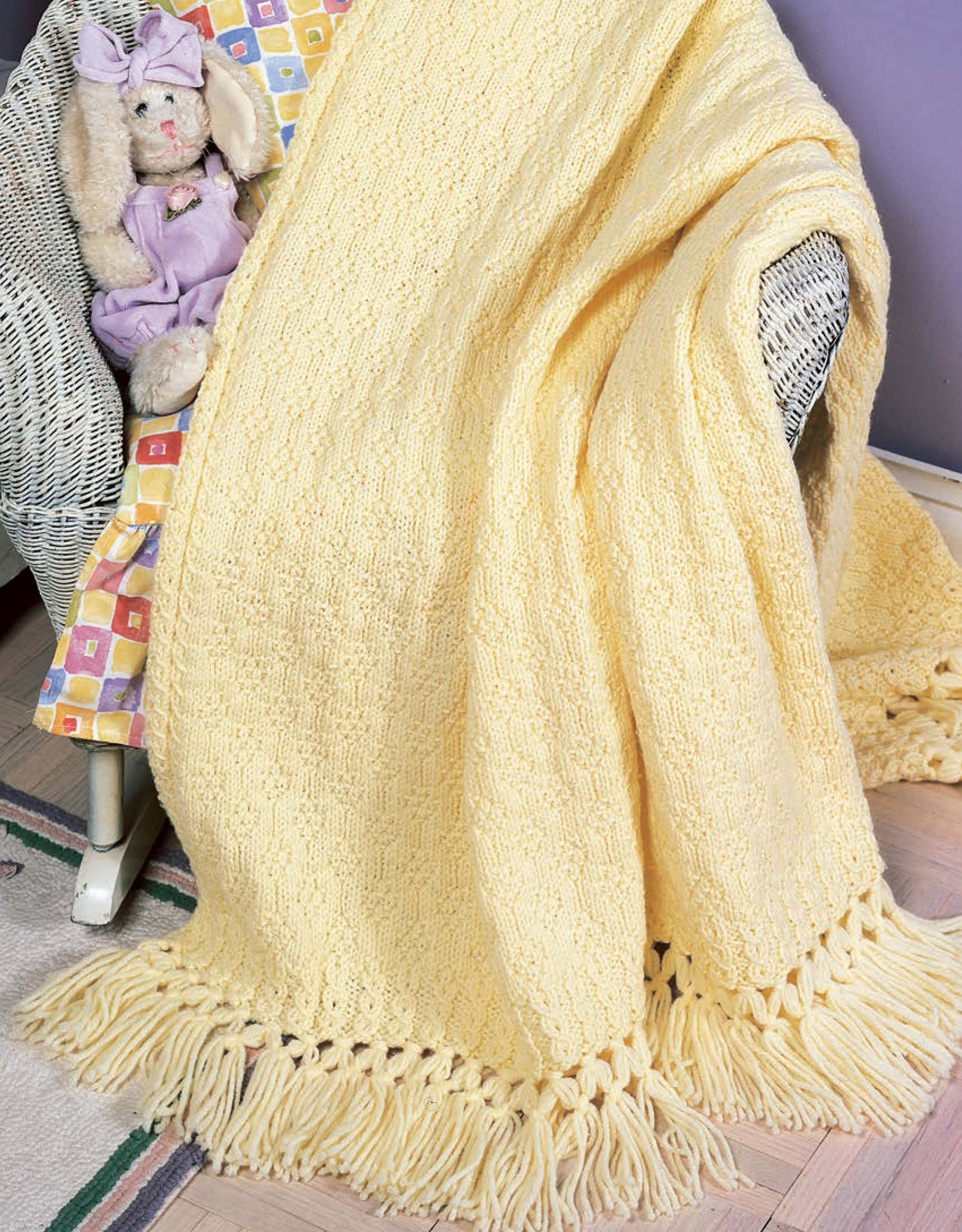 A cozy yellow blanket from Leisure Arts’ "Our Best Knit Baby Afghans" drapes over a white wicker chair, with a plush bunny in purple partially covered by the soft, fringed handmade knit.