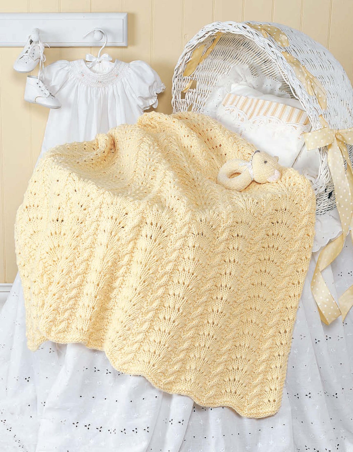 A soft yellow knitted baby blanket from "Our Best Knit Baby Afghans" by Leisure Arts drapes over a white bassinet with a stuffed bear and ruffled pillow, next to a white baby dress and hanging baby shoes.