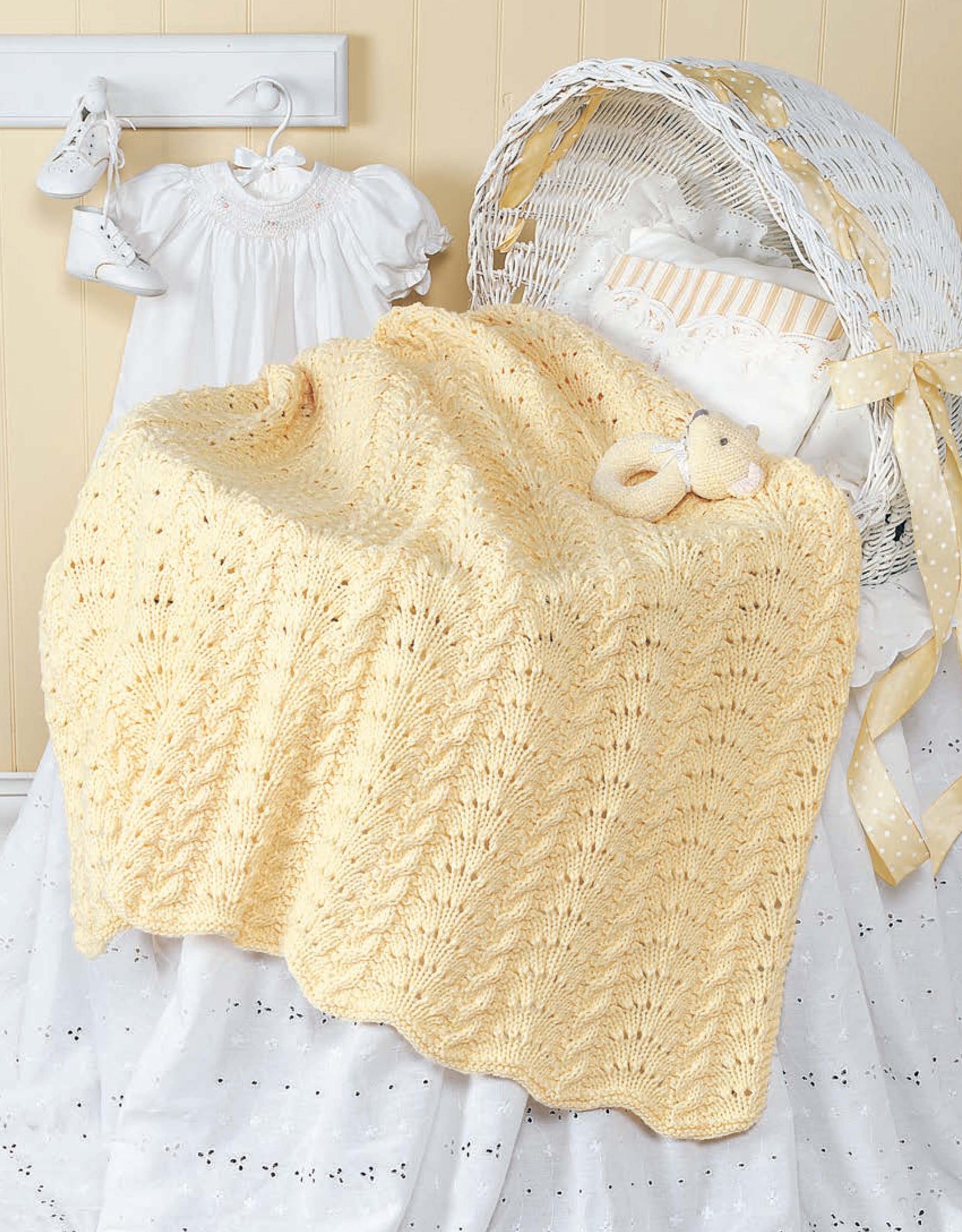 A soft yellow knitted baby blanket from "Our Best Knit Baby Afghans" by Leisure Arts drapes over a white bassinet with a stuffed bear and ruffled pillow, next to a white baby dress and hanging baby shoes.