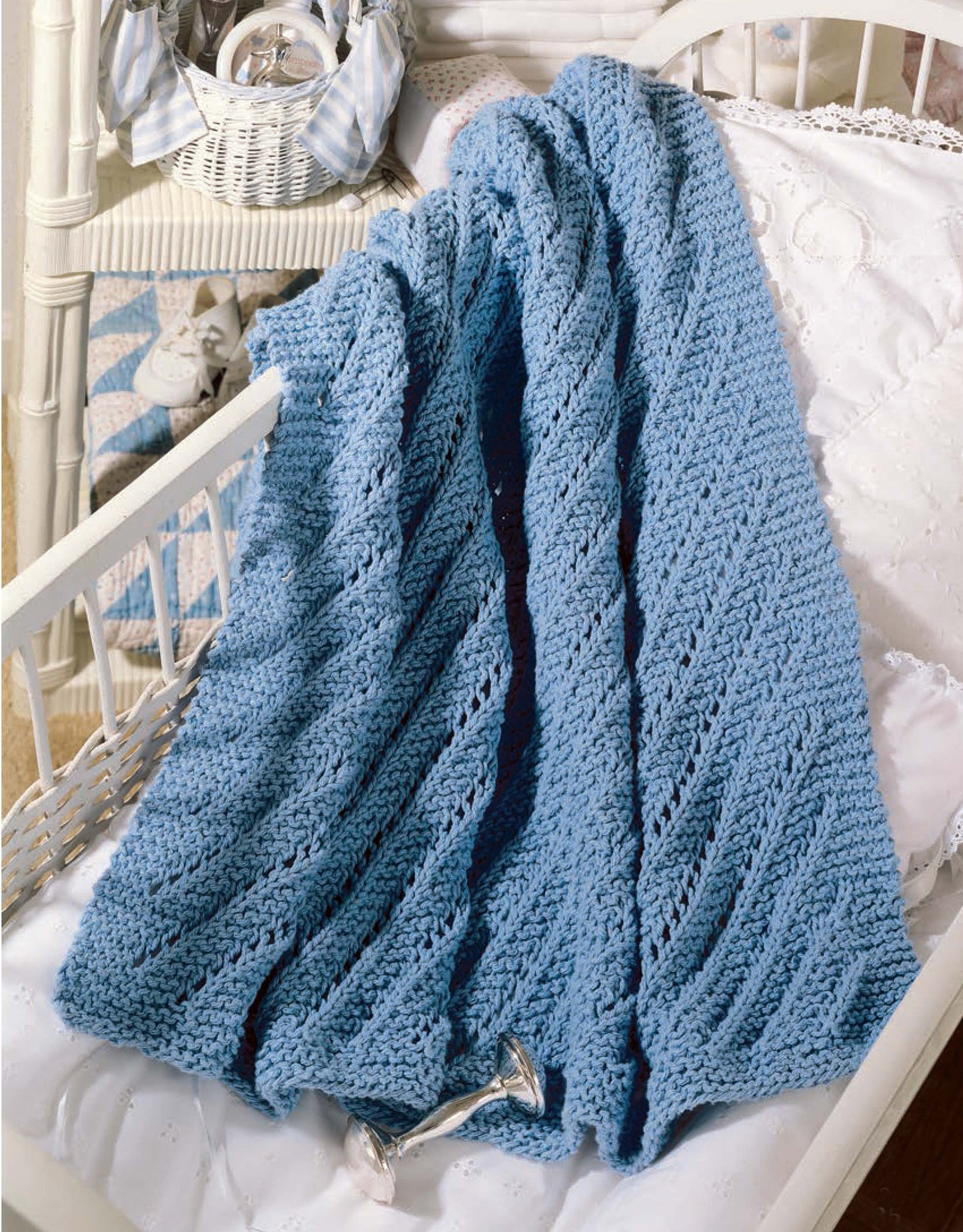 A light blue blanket made from Leisure Arts' "Our Best Knit Baby Afghans" drapes over a white crib with a rattle, adding soft charm to a cozy, light-decor nursery.