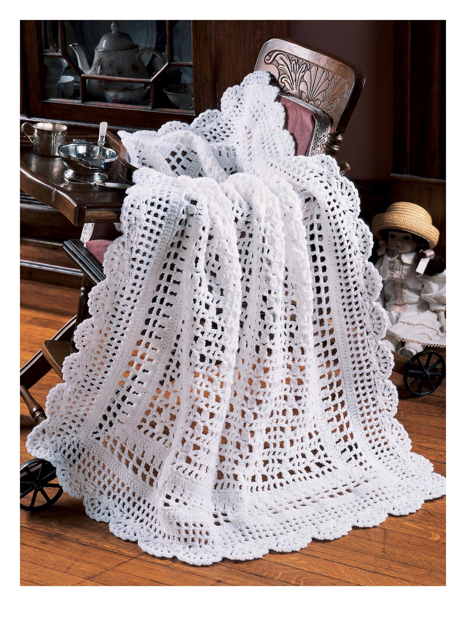 A delicate baby afghan from "Best of Terry Kimbrough Baby Afghans" by Leisure Arts drapes over a vintage chair beside a tea set, with a porcelain doll in a stroller nearby, evoking timeless designs on the wooden floor.