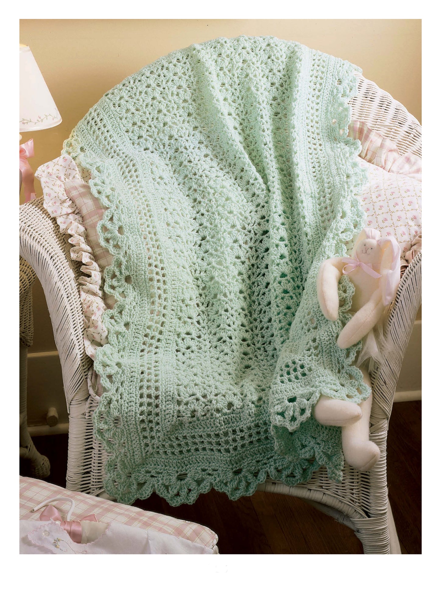 A light green crocheted baby blanket with a scalloped edge drapes over a white wicker chair, beside a stuffed bunny and patterned pillows—the perfect cozy setting to showcase classic afghans from Best of Terry Kimbrough Baby Afghans by Leisure Arts.