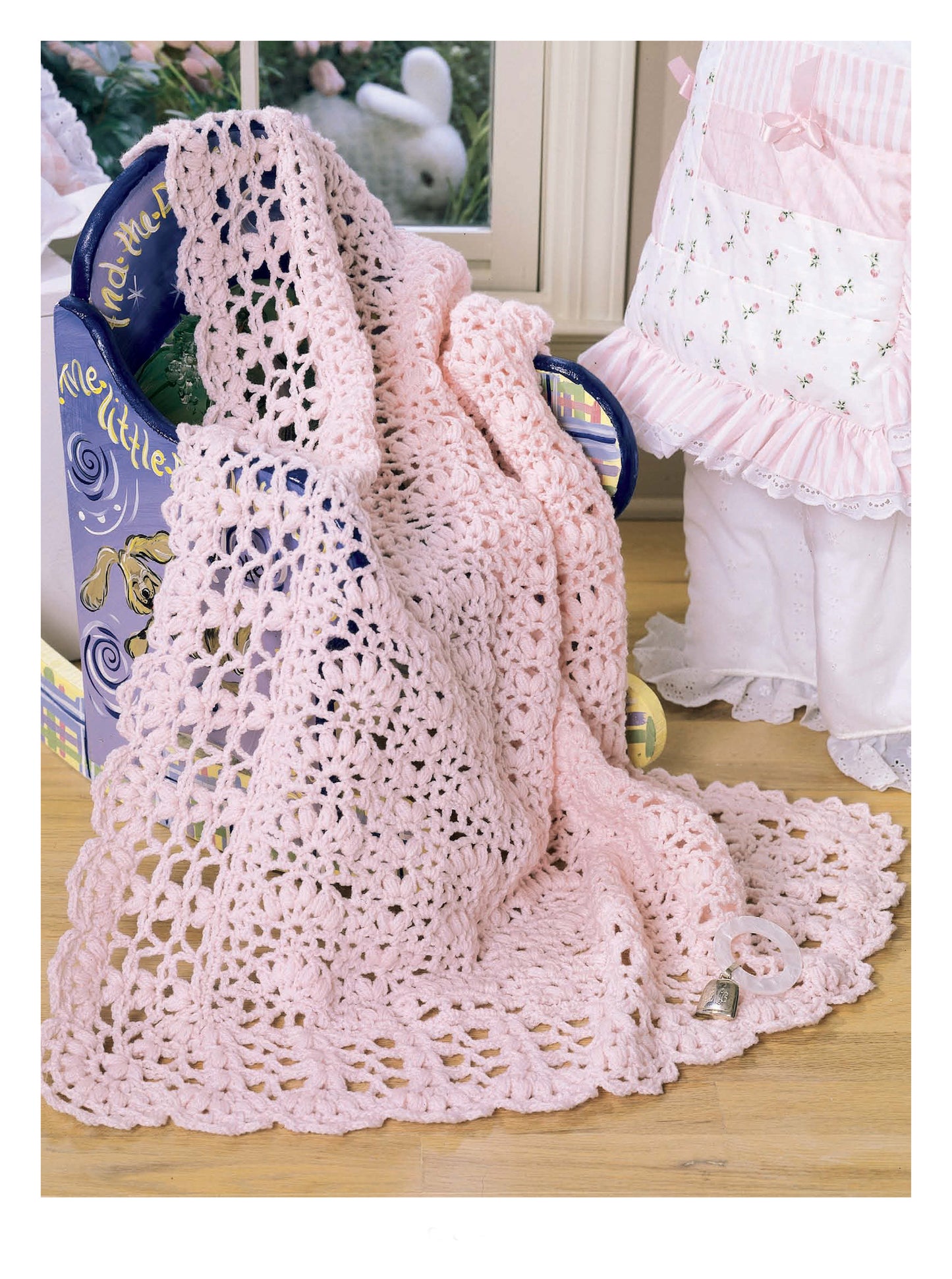 A soft, light pink crocheted baby blanket from the Leisure Arts "Best of Terry Kimbrough Baby Afghans" collection is draped over a blue chair with a doll, box, and baby outfit nearby; a pacifier rests on the wooden floor in front.
