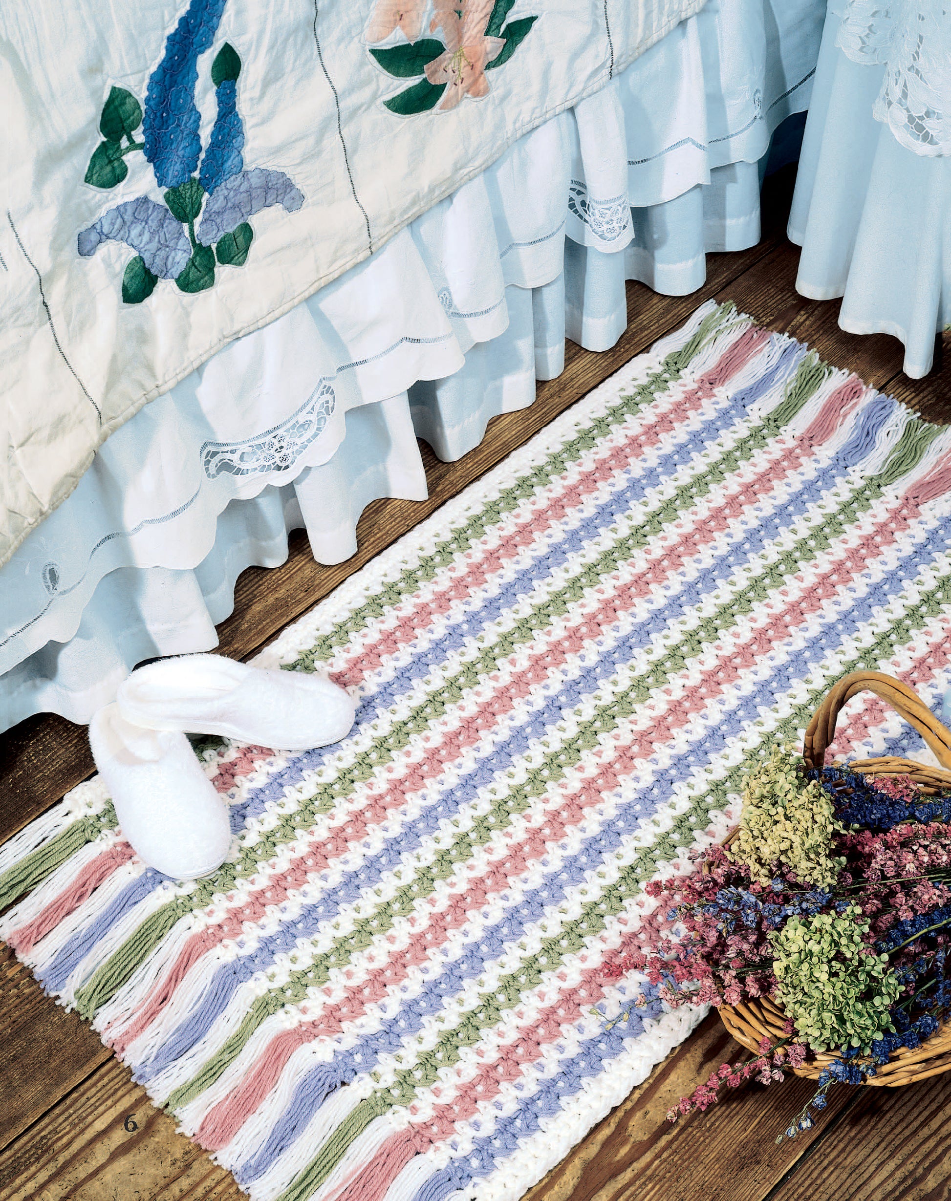 A striped, multicolored rug made with beginner techniques from "Easy Rugs to Crochet" by Leisure Arts lies on a wooden floor beside a bed with a ruffled embroidered quilt. White slippers rest on the rug and a basket of dried flowers sits nearby.