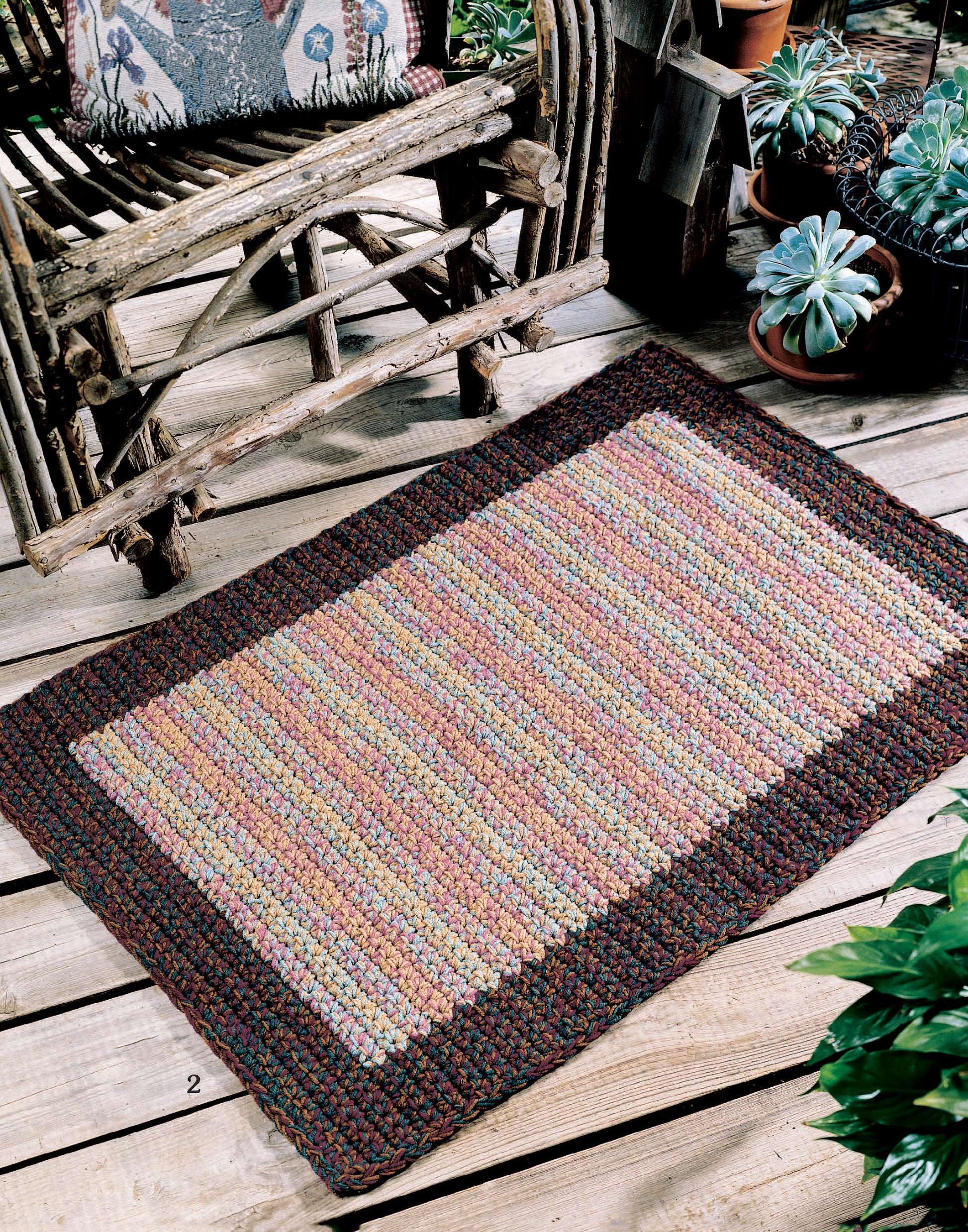 The "Easy Rugs to Crochet" by Leisure Arts features a rectangular woven rug with a multicolored striped center and dark border, placed on a wooden floor amid rustic furniture and potted succulents.