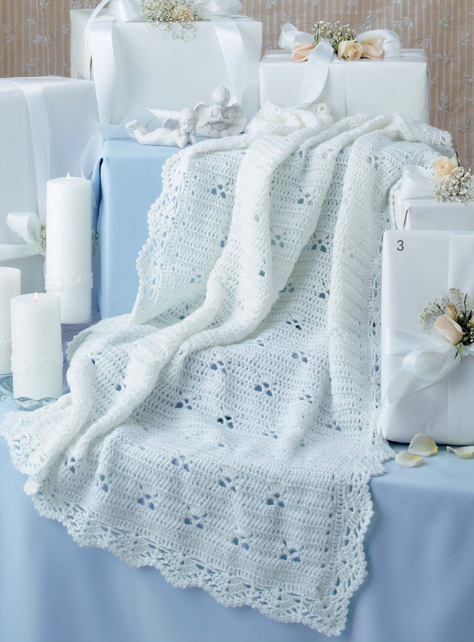 The Leisure Arts Keepsake Baby Afghans is a delicate white crocheted blanket displayed on a blue table, surrounded by ribboned gift boxes, candles, and soft floral accents, creating an elegant scene with timeless charm.
