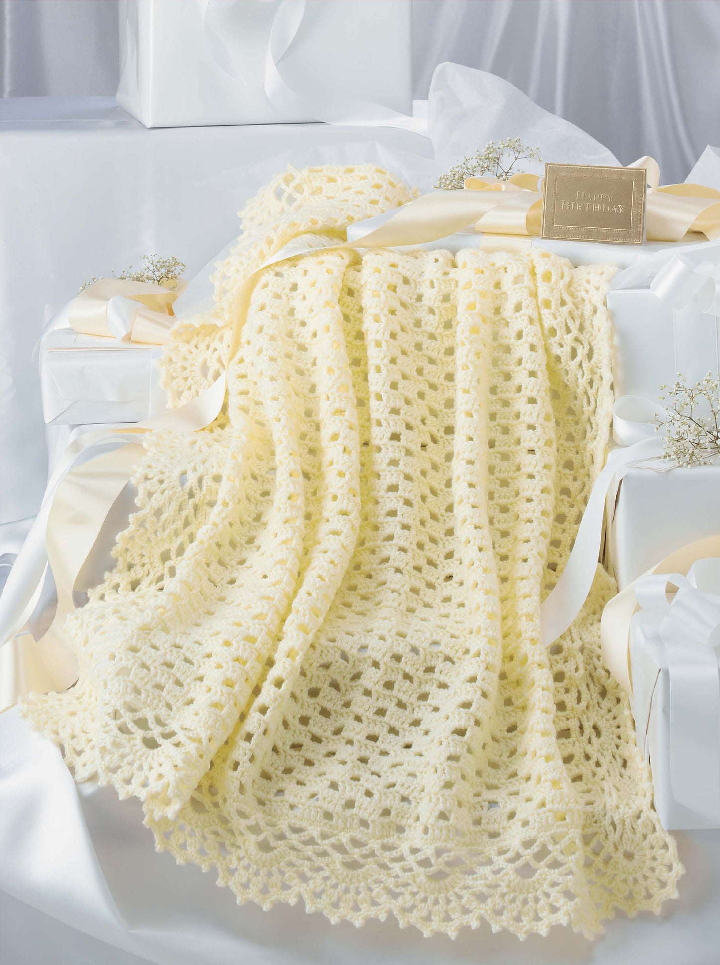 The Leisure Arts Keepsake Baby Afghans, a soft pale yellow crocheted blanket with elegant edgings, is draped over white gift boxes with cream ribbons, displayed on white satin beside baby's breath and a gold card.