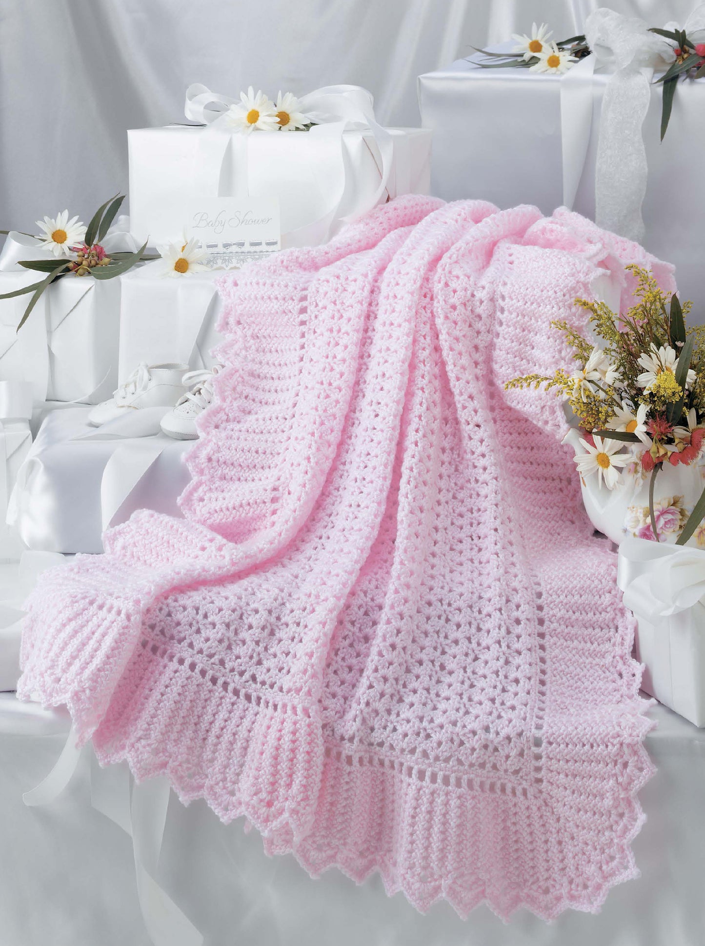 The Leisure Arts Keepsake Baby Afghans, in soft pink with elegant edgings, is displayed among white gift boxes and daisies, adding vintage charm and festive elegance to the scene.