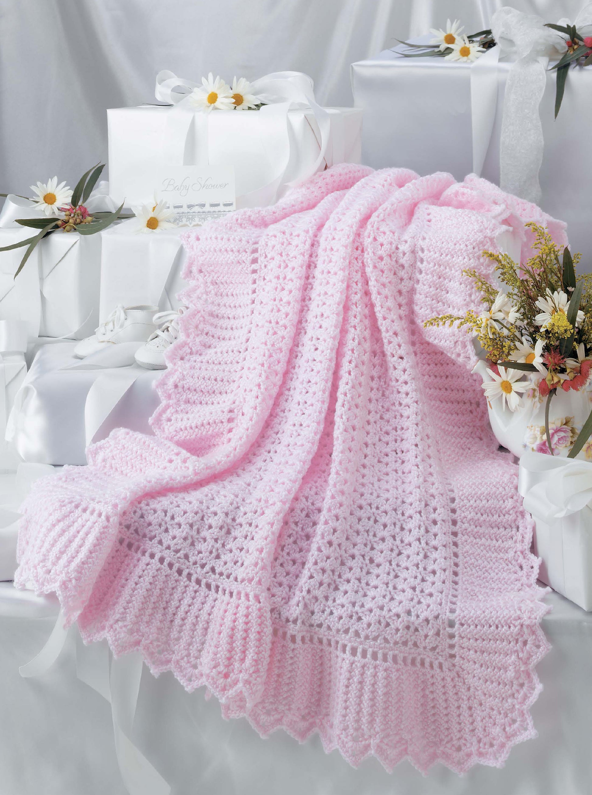 The Leisure Arts Keepsake Baby Afghans, in soft pink with elegant edgings, is displayed among white gift boxes and daisies, adding vintage charm and festive elegance to the scene.