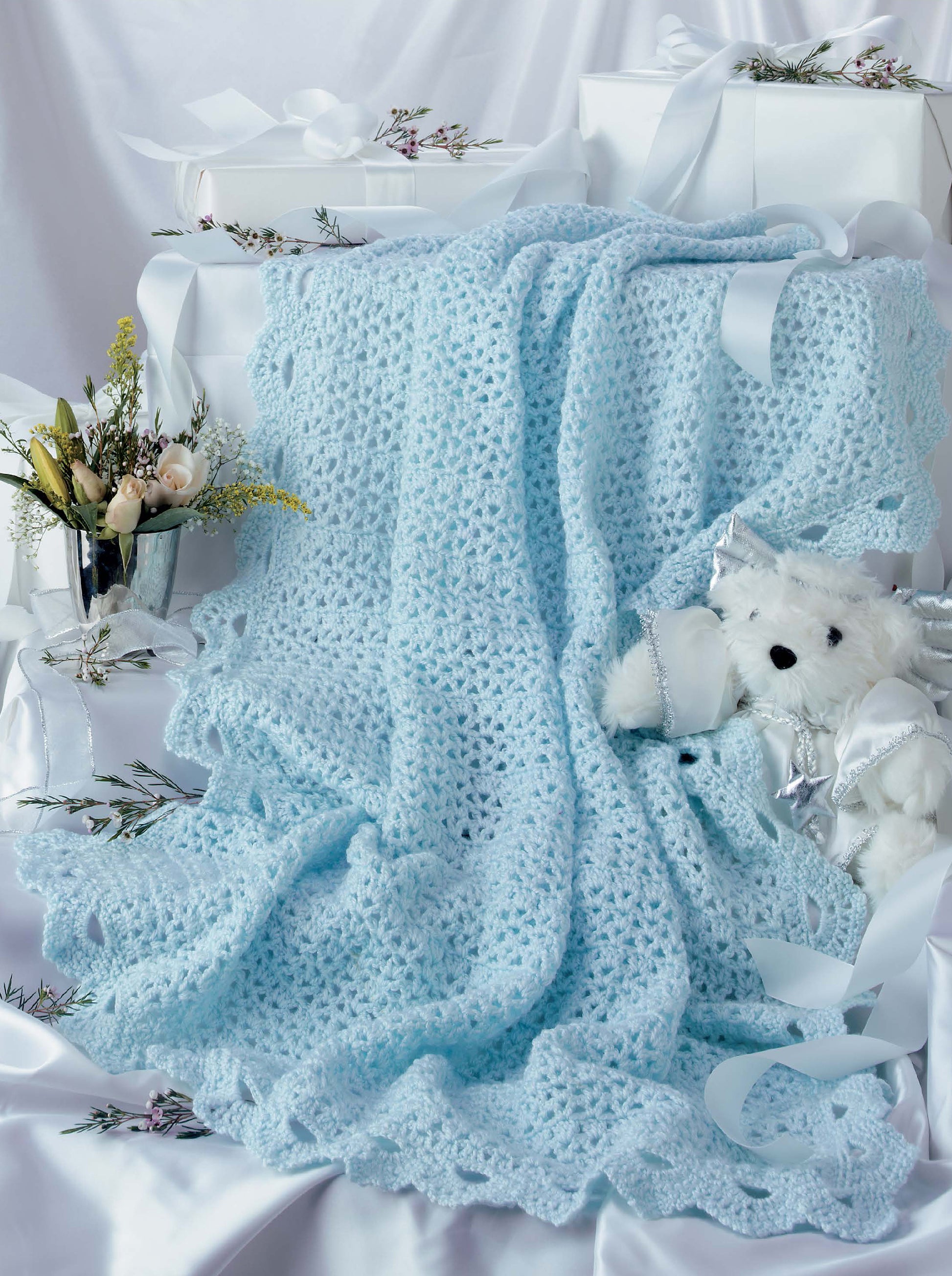 The Leisure Arts Keepsake Baby Afghans features a soft blue crocheted blanket with elegant edgings, displayed with white gift boxes, flowers, and a plush teddy bear—ideal for crochet lovers who cherish delicate details.