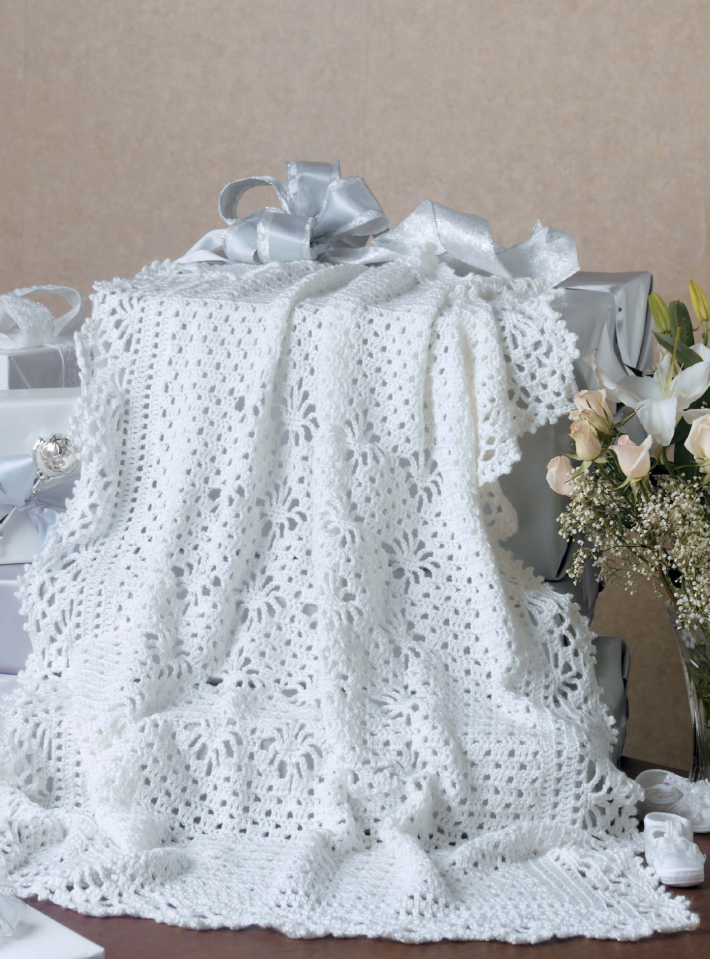 The Leisure Arts Keepsake Baby Afghans, a white crocheted blanket with floral patterns and elegant edgings, is draped over silver-wrapped gift boxes beside a bouquet of white roses and baby's breath on a neutral background.