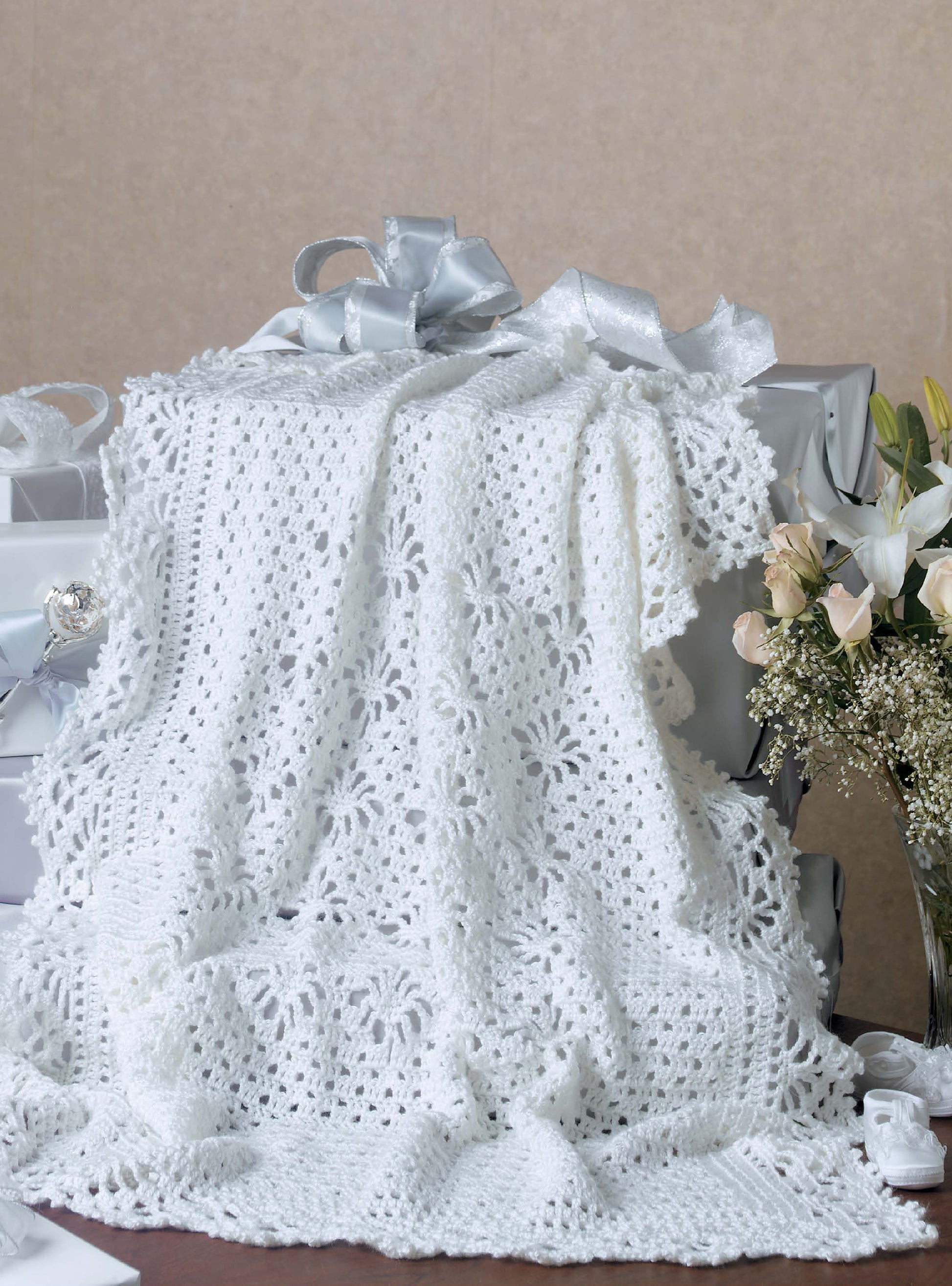 The Leisure Arts Keepsake Baby Afghans, a white crocheted blanket with floral patterns and elegant edgings, is draped over silver-wrapped gift boxes beside a bouquet of white roses and baby's breath on a neutral background.
