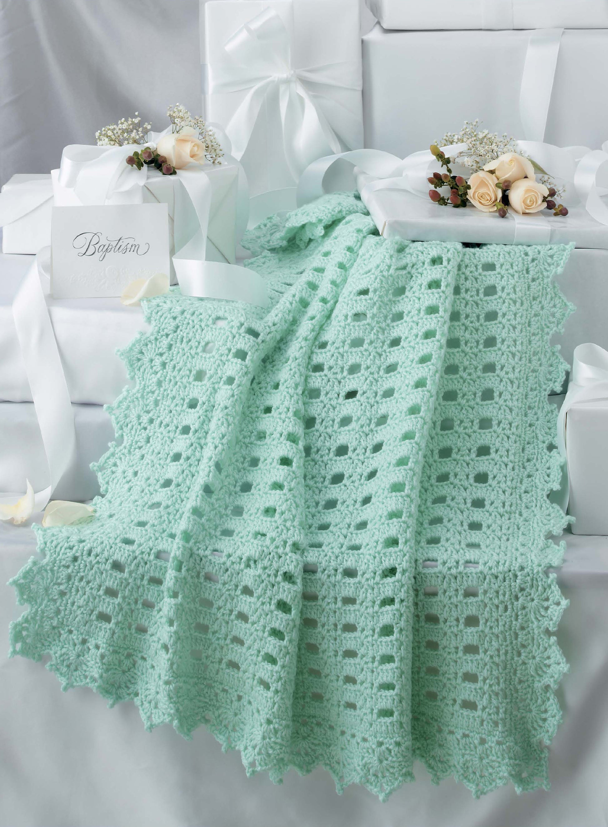 The Keepsake Baby Afghans by Leisure Arts, in mint green with elegant edging and vintage charm, is displayed alongside white satin-tied gift boxes, ivory roses, baby's breath, and a Baptism card.