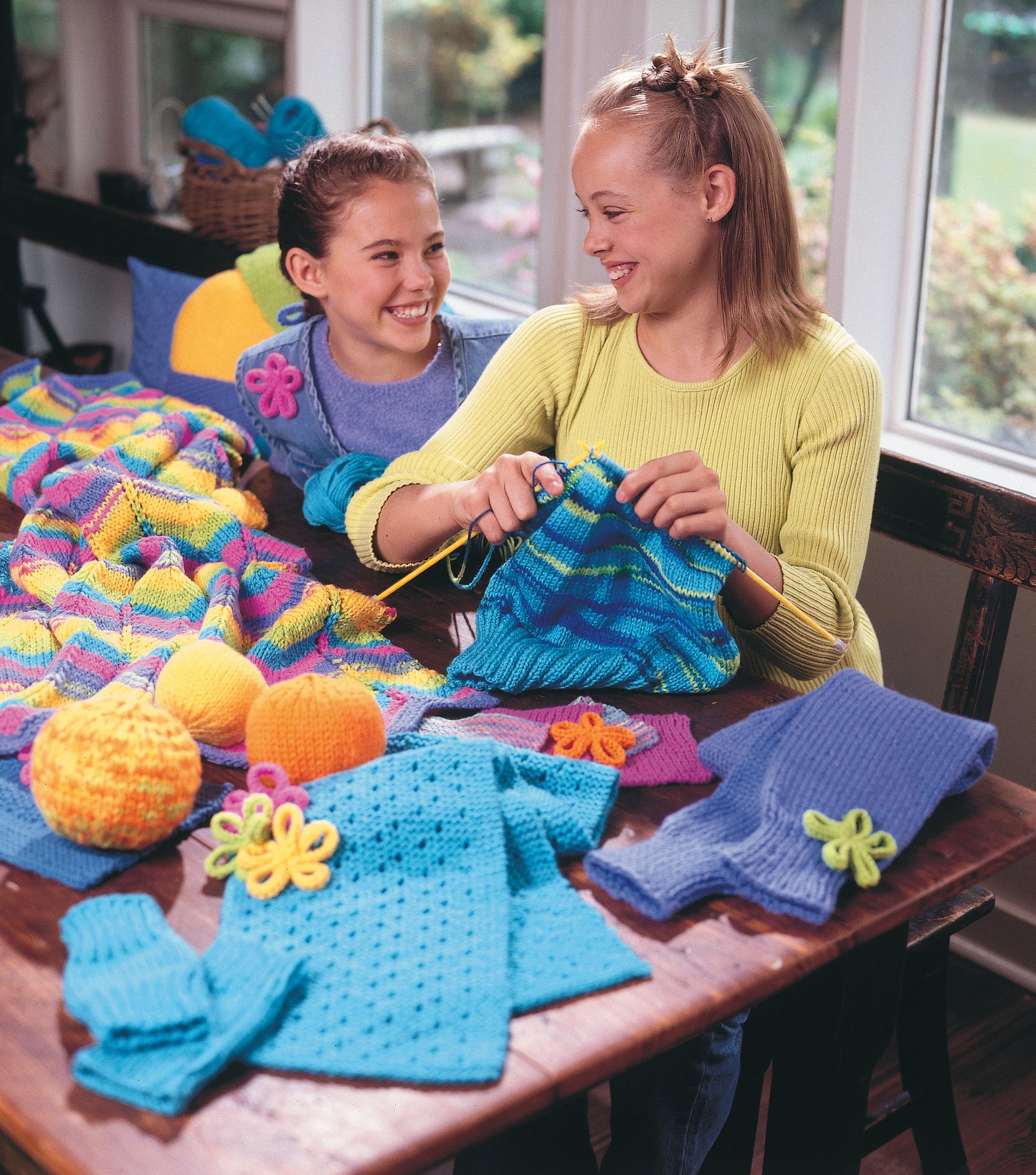 Two girls, smiling and knitting together, sit at a table covered with bright sweaters, yarn balls, and Leisure Arts' Cool Stuff for Kids Teach Me to Knit—12 Projects in a cozy room filled with sunlight.